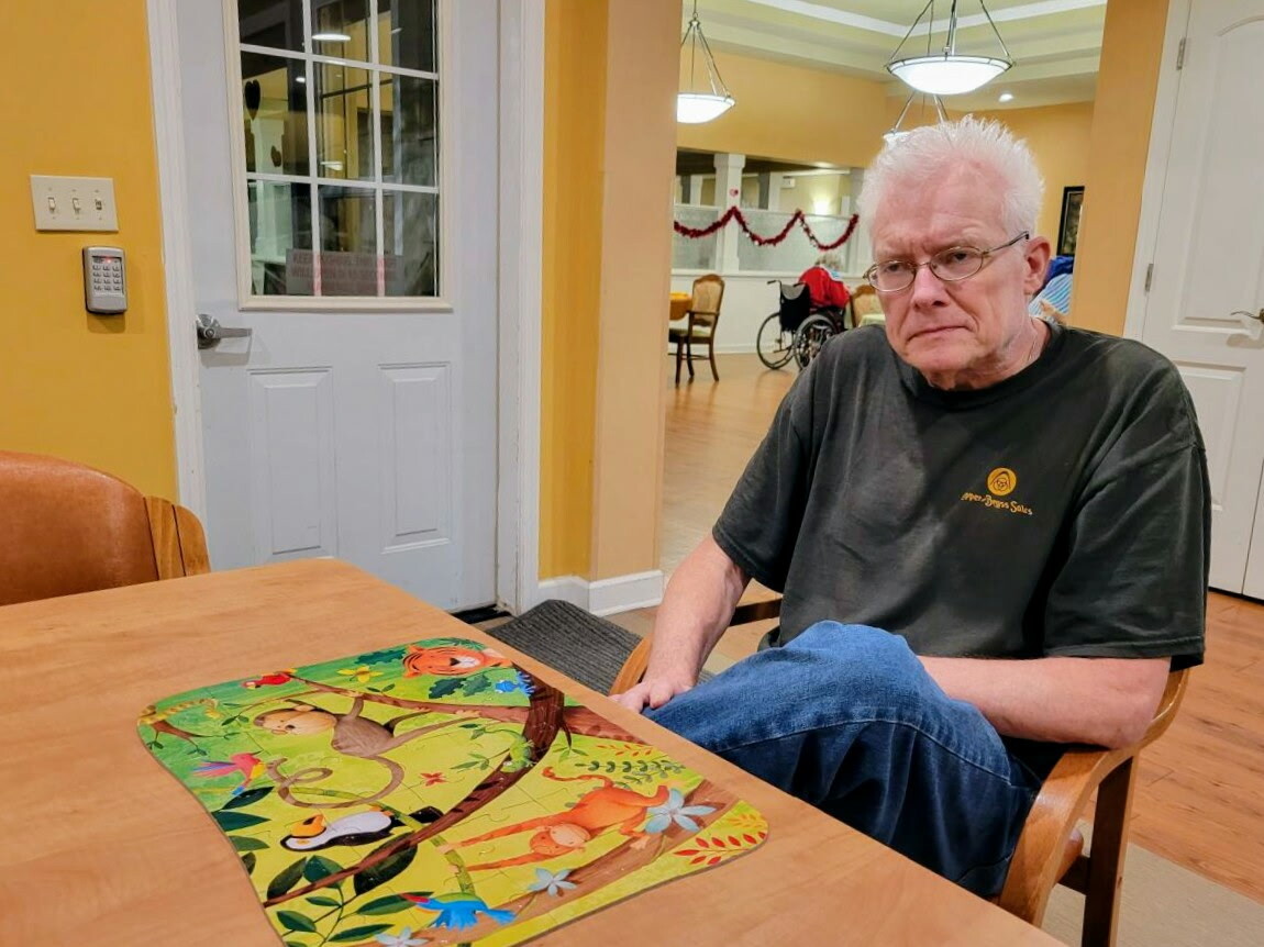 Older man with white hair and glasses sits at a wooden table in a yellow-walled room, a colorful animal-themed puzzle on the table in front of him.