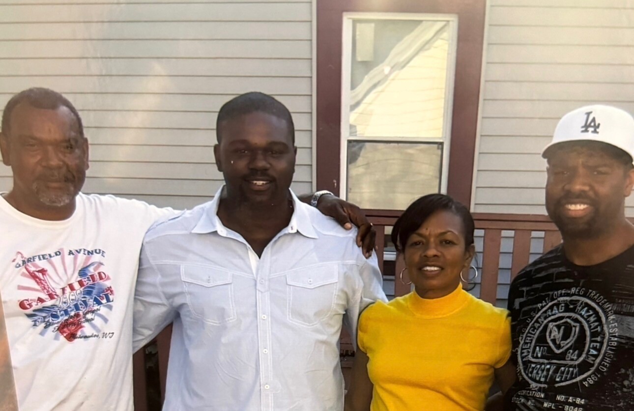 Four adults stand together on a porch, smiling at the camera; from left, a man in a white graphic tee, a man in a light blue button‑down shirt, a woman in a yellow top, and a man in a black shirt and white cap.