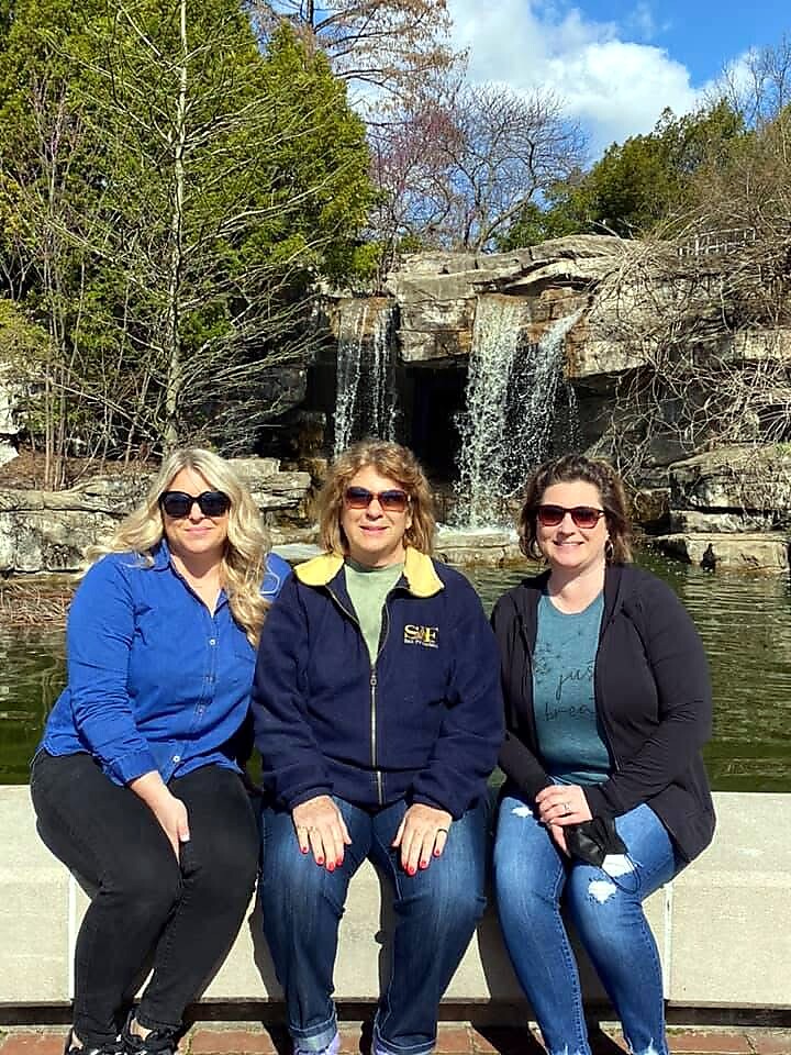 Three women sit on a low wall in front of a waterfall and pond, posing for a sunny outdoor photo.