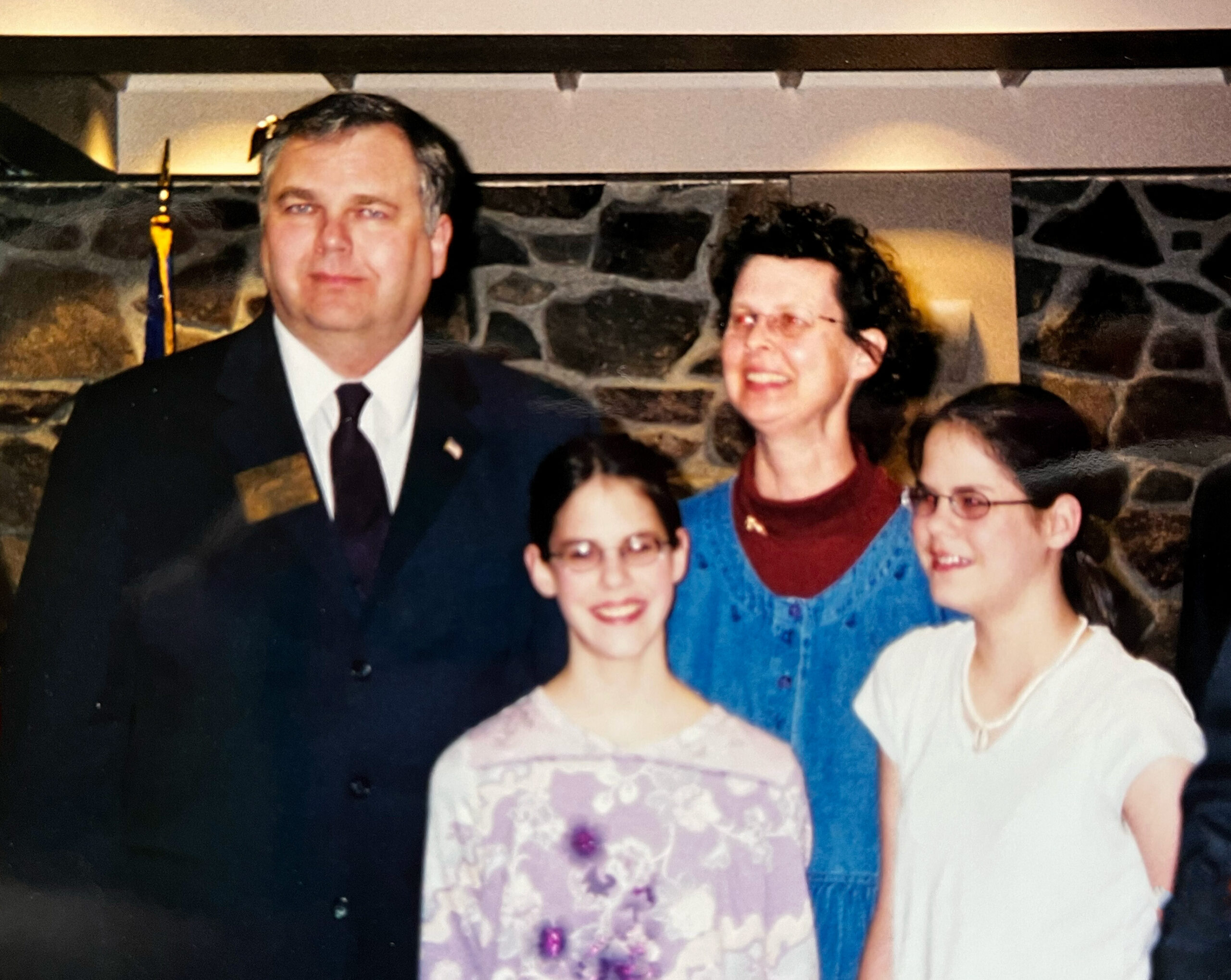 Group portrait at a formal event: man in suit on the left, smiling family members surrounding him against a stone wall backdrop.