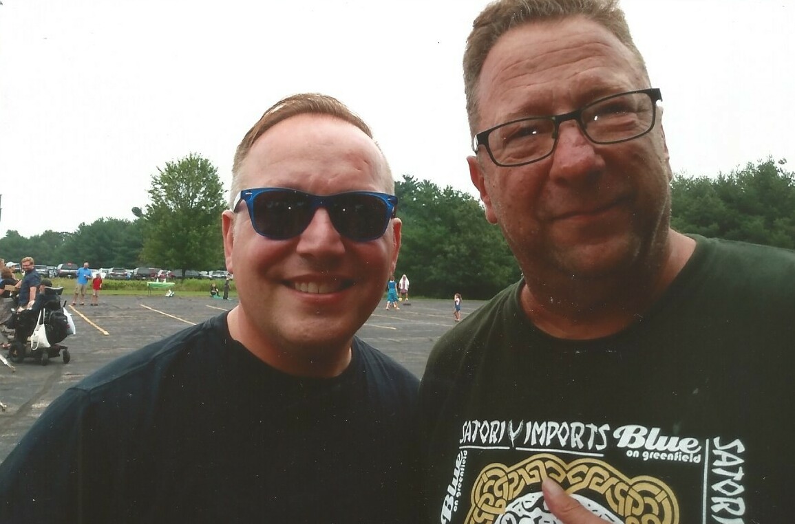 Two men posing for a selfie in a parking lot, smiling; one wears sunglasses and a black shirt, the other in a green shirt with graphic print.