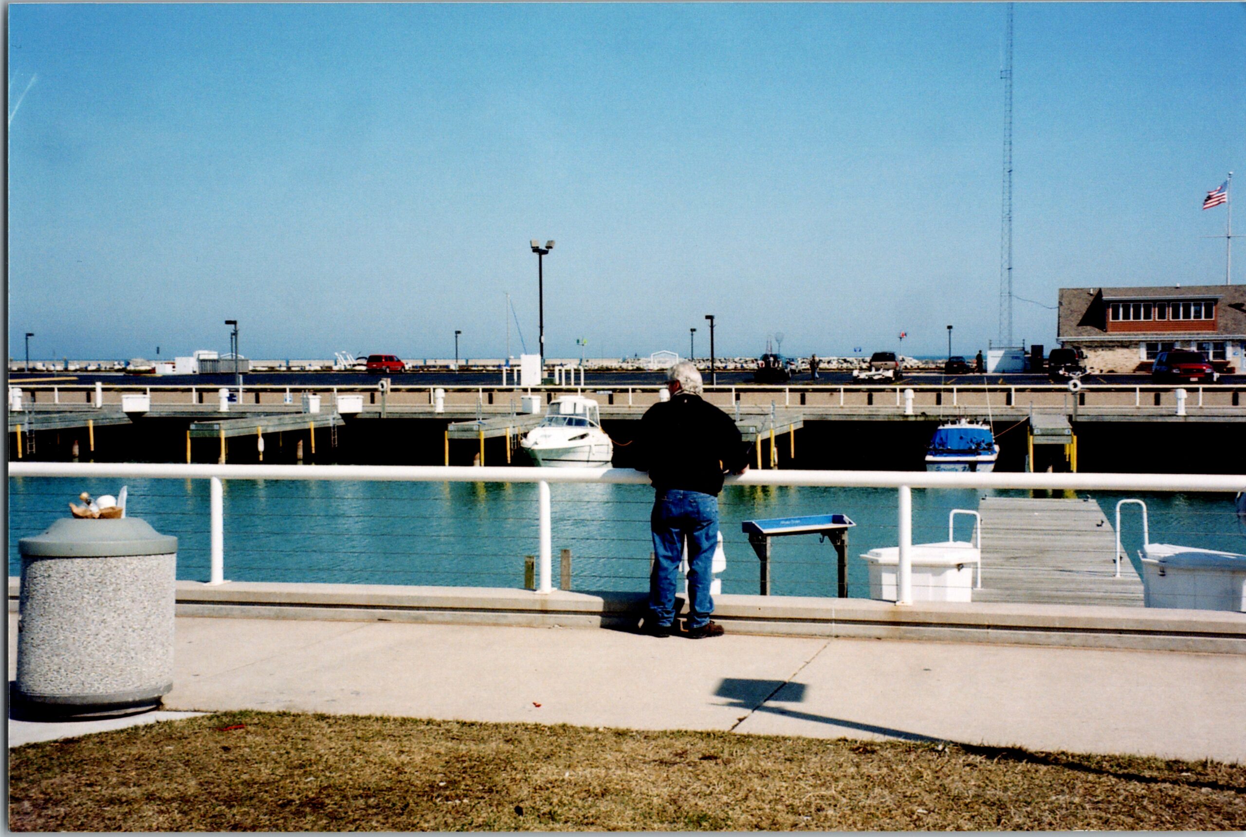 Man with a dark jacket and jeans stands on a marina dock facing the water, with boats moored behind him under a blue sky.