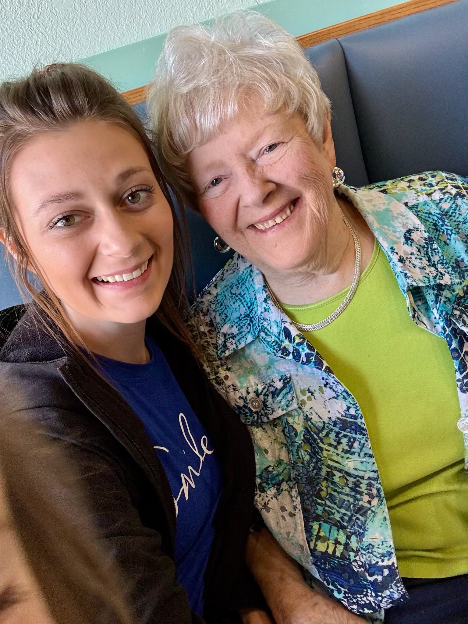 Two women smiling for a selfie, one younger on the left and an older woman on the right wearing a green top and patterned jacket.