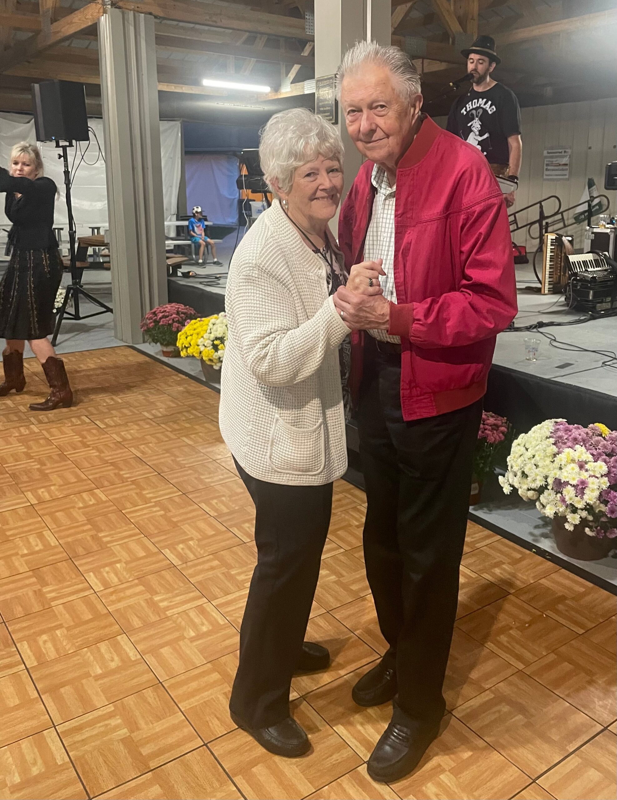 Older couple dancing in a hall at a community event; woman with white hair in a light cardigan and man in a red jacket hold hands as they dance, stage with a performer and instruments in the background, and potted flowers nearby.