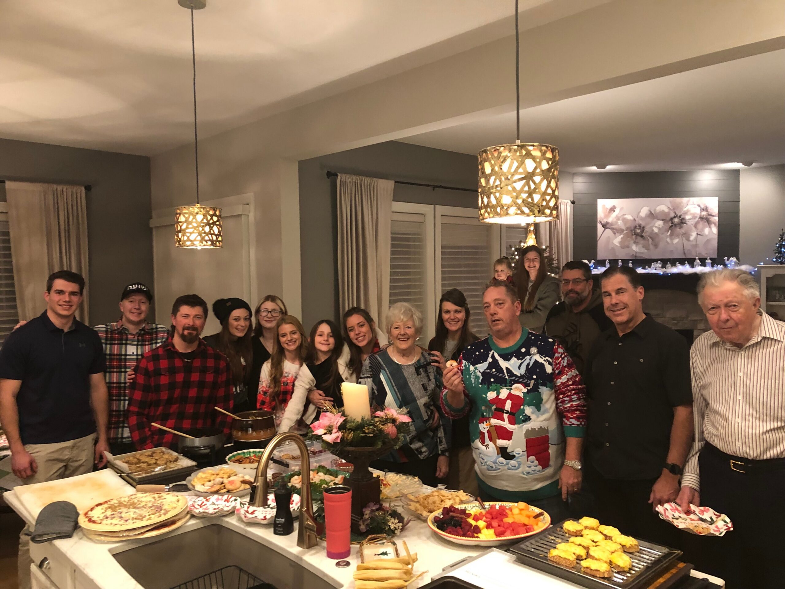 Group of people gathered around a holiday buffet in a kitchen, smiling at the camera. Front table has pies, fruit and snacks, with warm pendant lights overhead.