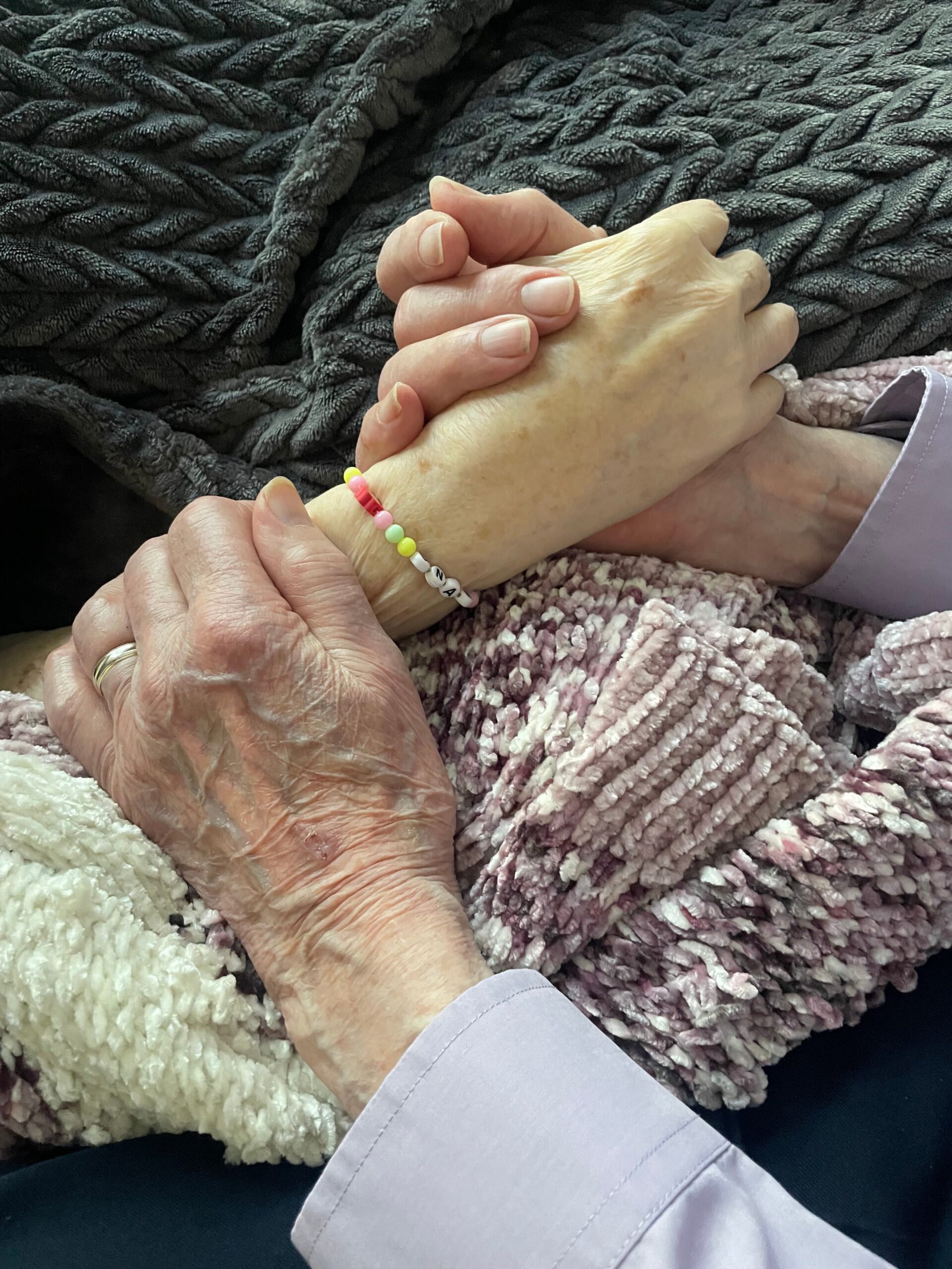 Two elderly hands clasped together on a soft textured blanket, wearing a colorful bead bracelet.