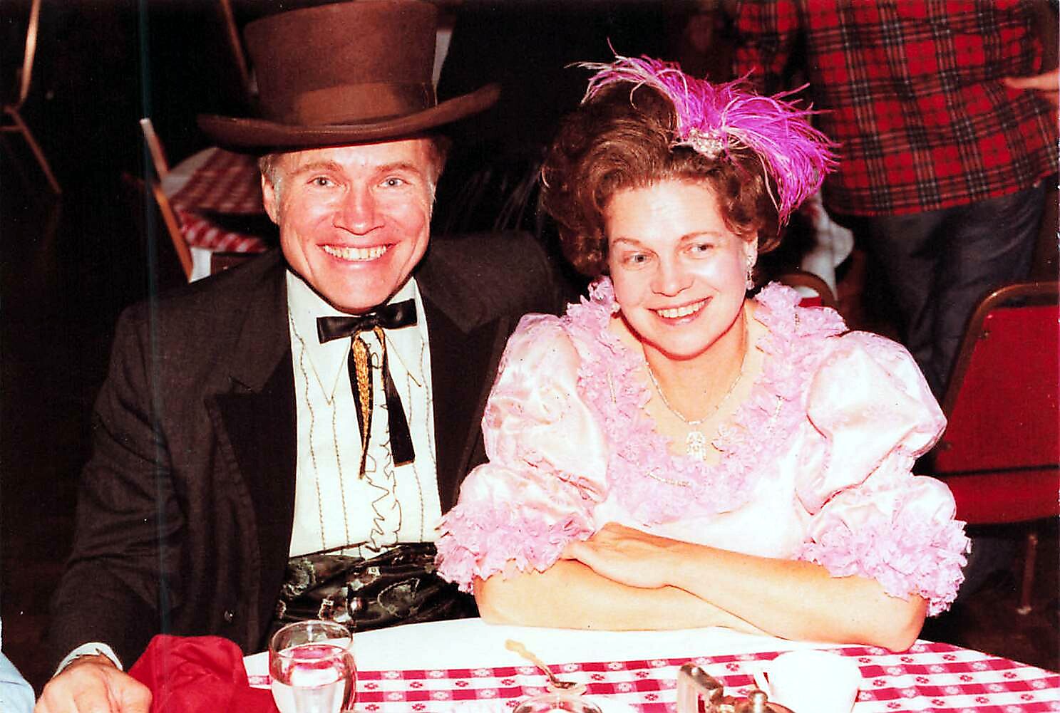 Smiling couple at a festive event: man in a top hat and bow tie, woman in a pink, ruffled dress with a pink feather headpiece, seated at a checkered tablecloth.