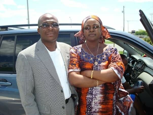 Man in a gray checkered suit jacket and sunglasses stands beside a woman in a colorful traditional orange-and-purple dress and headscarf, in front of a blue SUV with the door open.