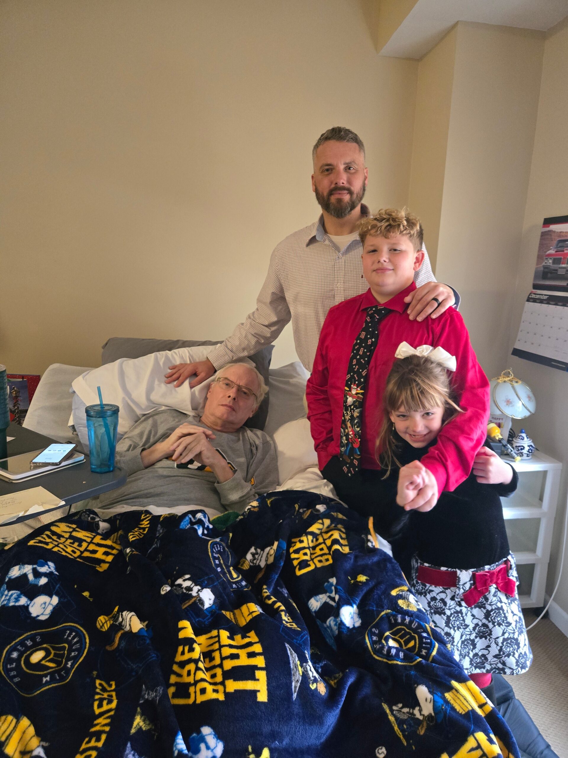 Family group photo: an adult man with a beard stands beside a hospital bed, with a boy in a red shirt and a girl with a bow posing beside him. An elderly man rests in bed under a dark blanket.