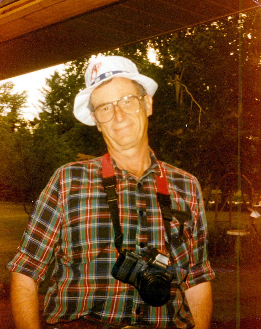 Older man in a white bucket hat and plaid shirt with a camera hanging from a red strap around his neck outdoors, smiling at the camera.