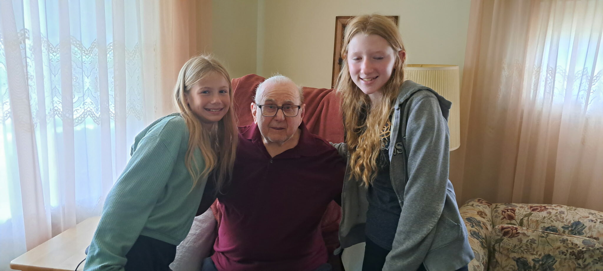 Two young girls smile and hug an elderly man seated on a chair in a cozy living room.