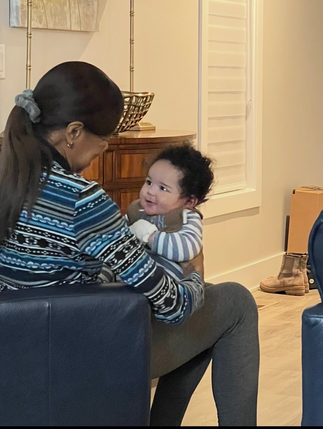 Woman in a blue patterned sweater sits on a chair, holding a smiling baby looking up at her indoors.