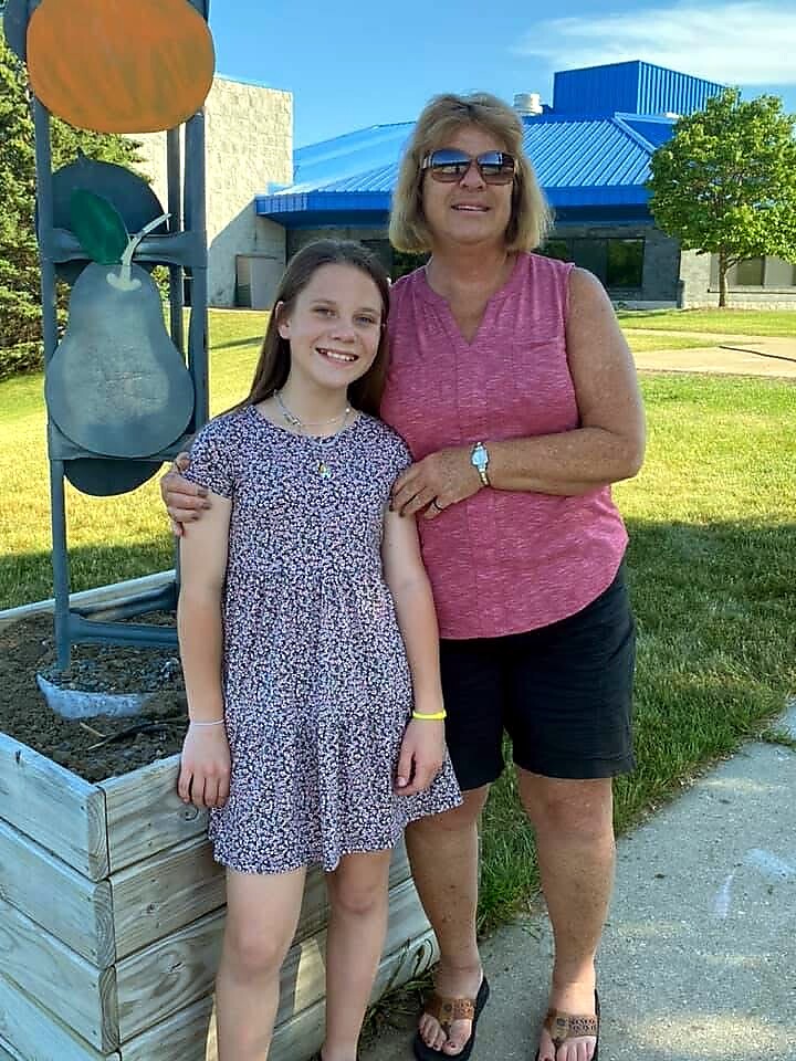 Grandmother and granddaughter posing together outdoors beside a wooden planter with a playground sculpture behind them.