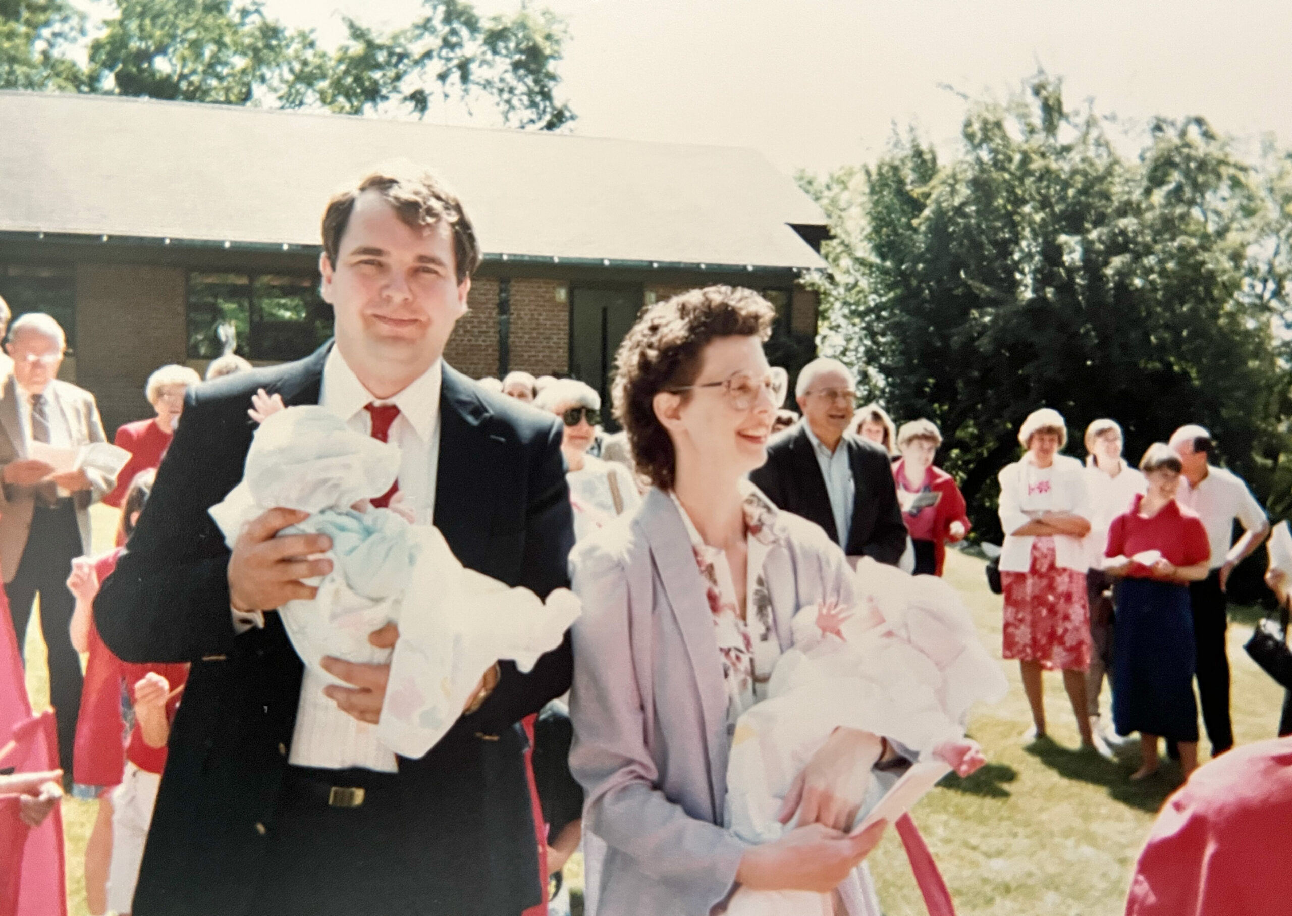Man in a dark suit holds a baby wrapped in a blanket; woman beside him smiles, with guests in bright clothing at an outdoor wedding.