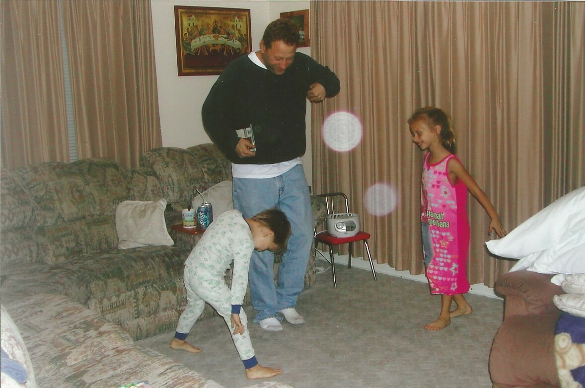 A man in a dark sweatshirt watches two children playing in a living room on a beige carpet.