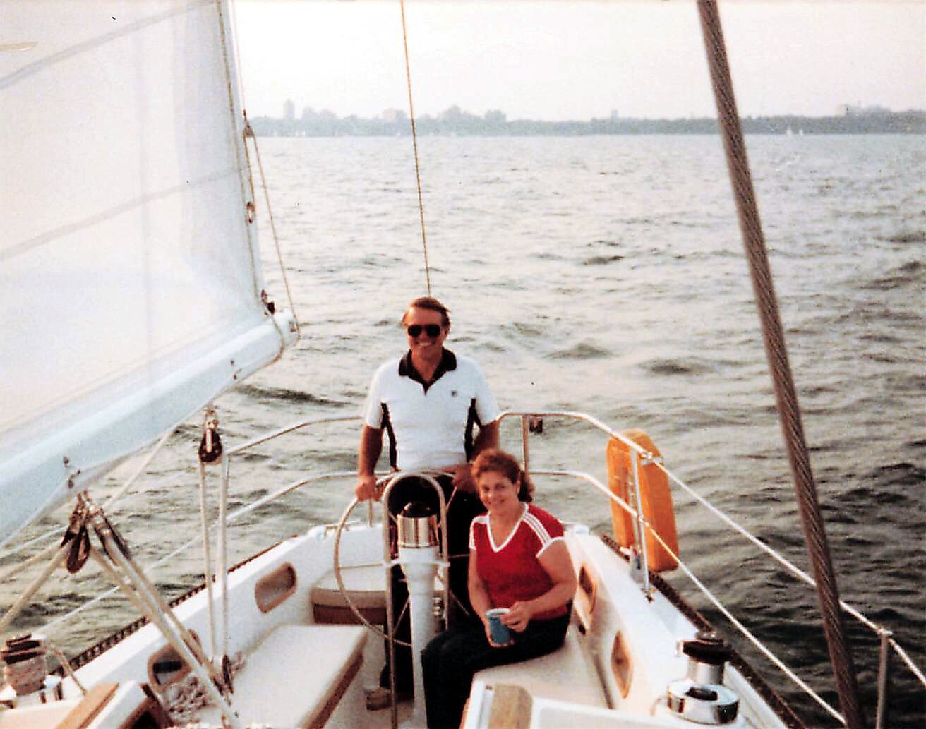 Man and woman on a sailboat steering near the cockpit, open water with a distant city skyline.