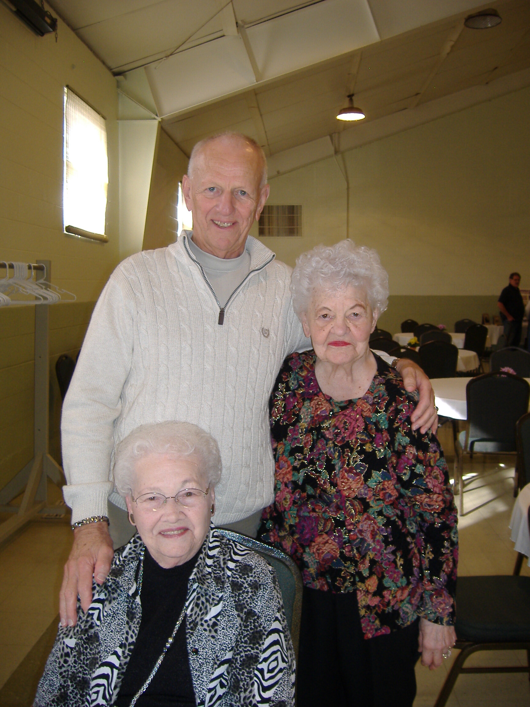 Group of four elderly people posing for a photo in a bright room with round tables in the background; man standing behind women, all smiling.