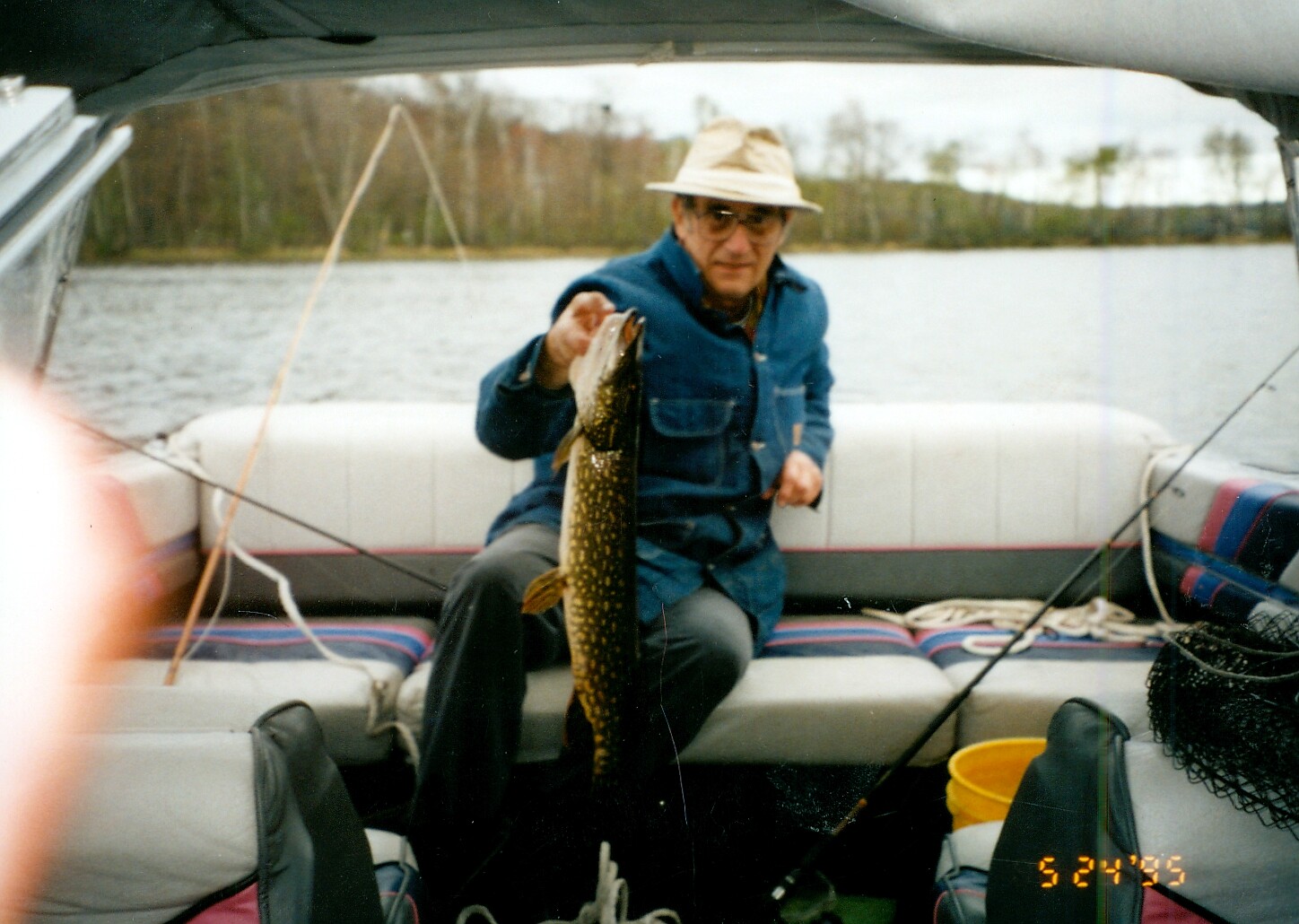 Older man in a beige hat holding a large fish on a small boat over a lake.