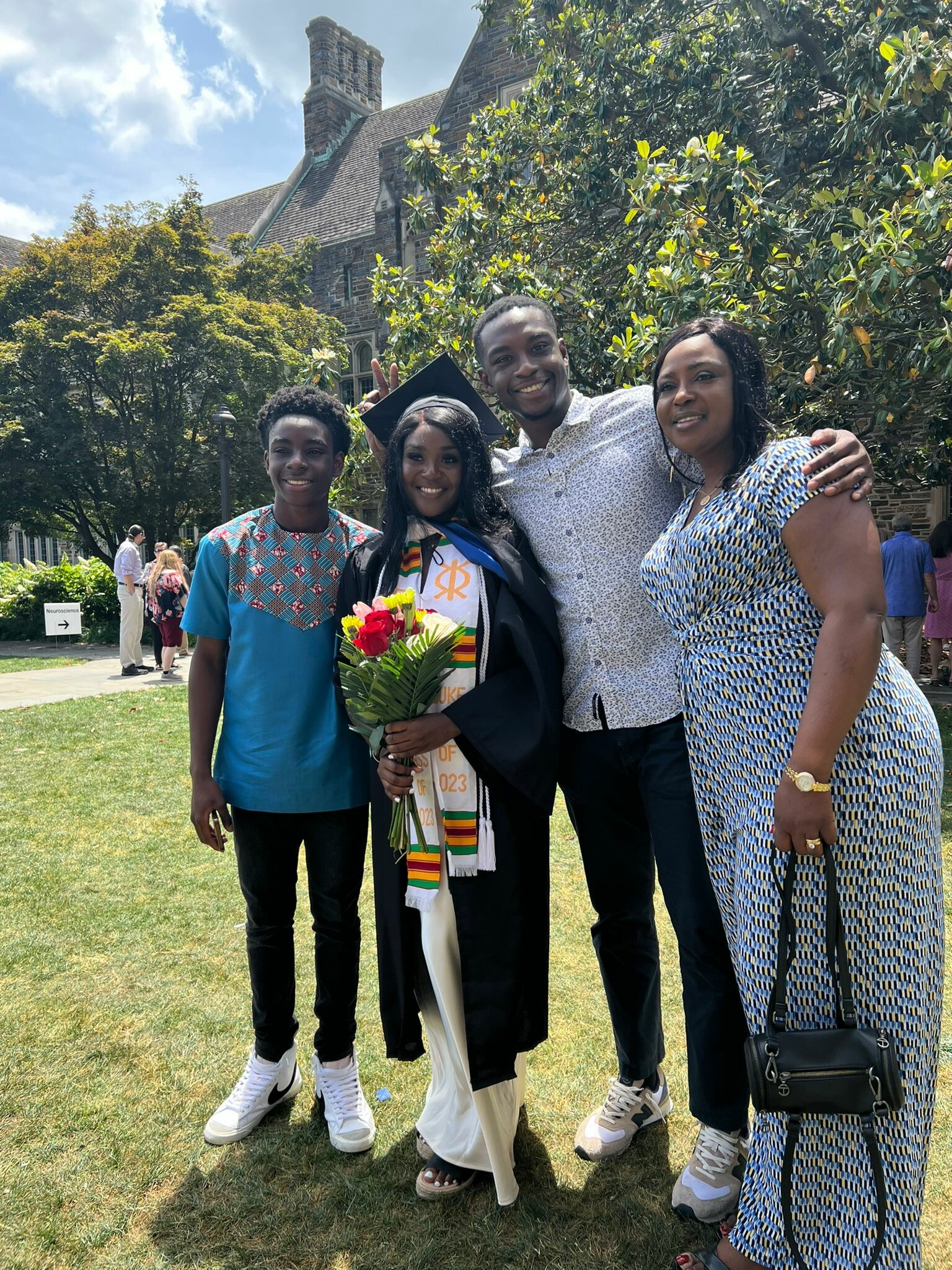 Group of four people posing outdoors on a sunny day, one graduate in cap and gown holding a bouquet with family members smiling beside them.