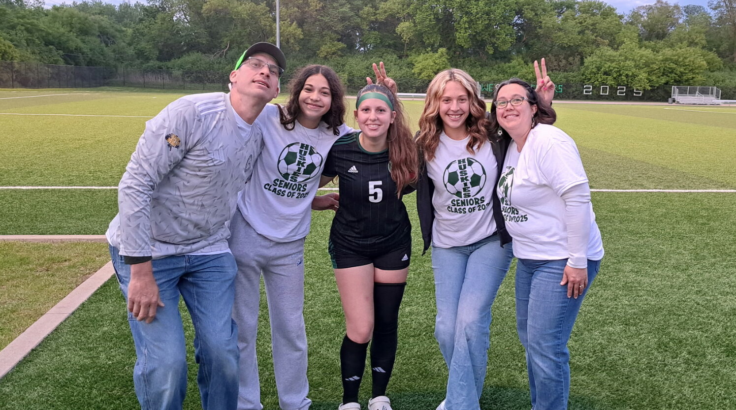 Group of five people posing on a soccer field; center player wears black uniform with number 5, others in white senior shirts.