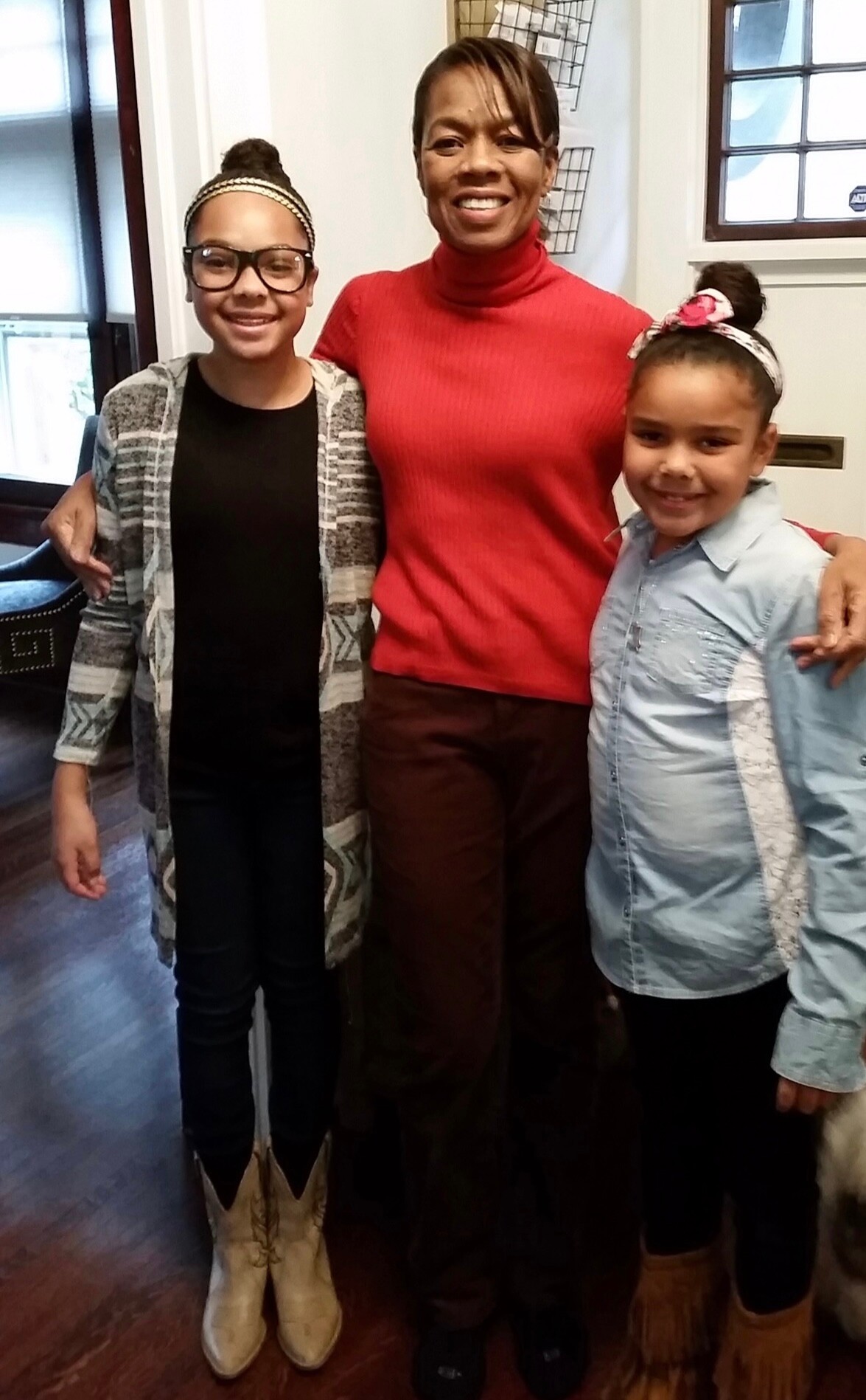 Smiling woman in a red turtleneck posing with two girls indoors, all smiling for a photo.
