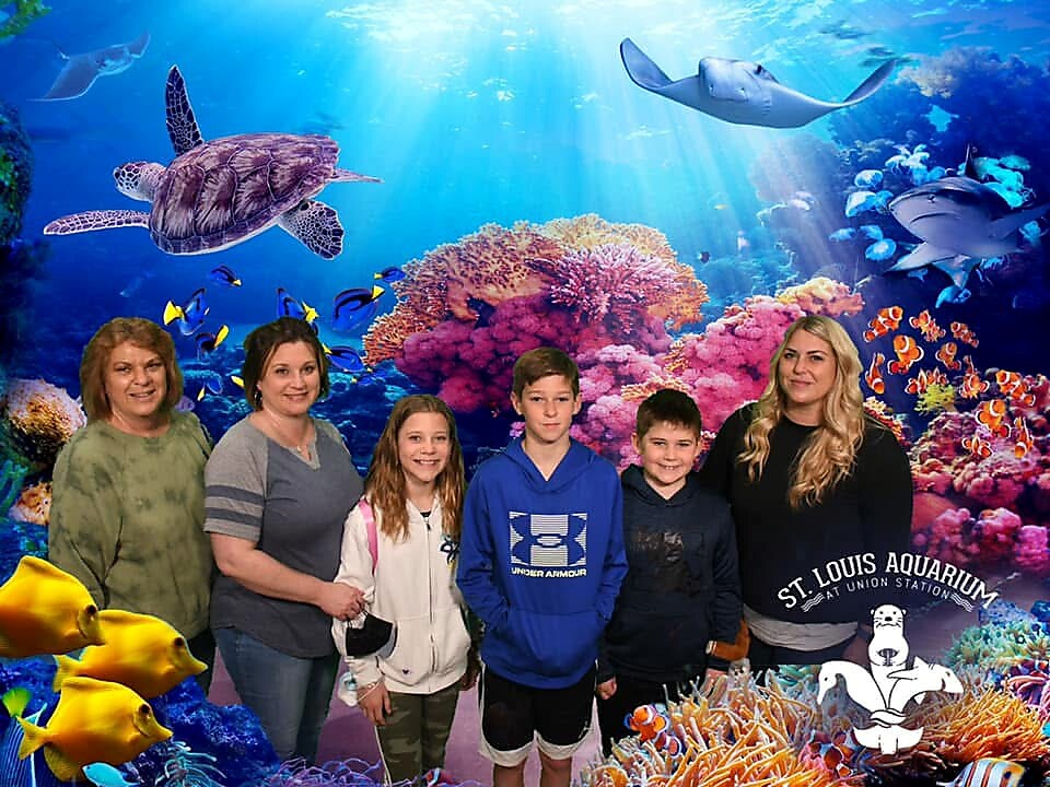 Six people stand together and smile in front of a vibrant coral reef mural with fish and sharks in an aquarium backdrop, logo bottom right.