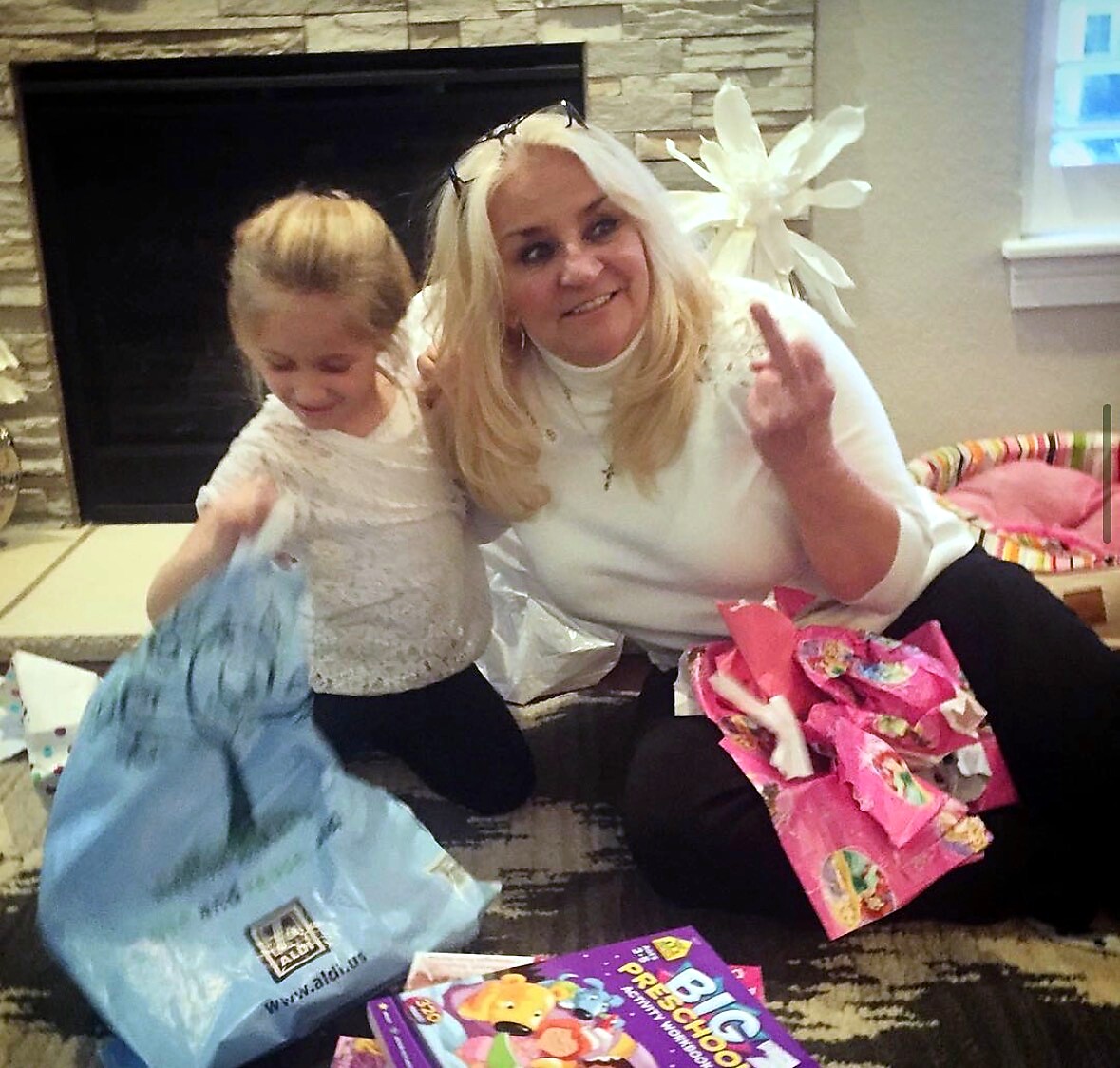 Smiling woman and young girl sit on the floor amid gifts and wrapping paper by a stone fireplace, posing for a photo.