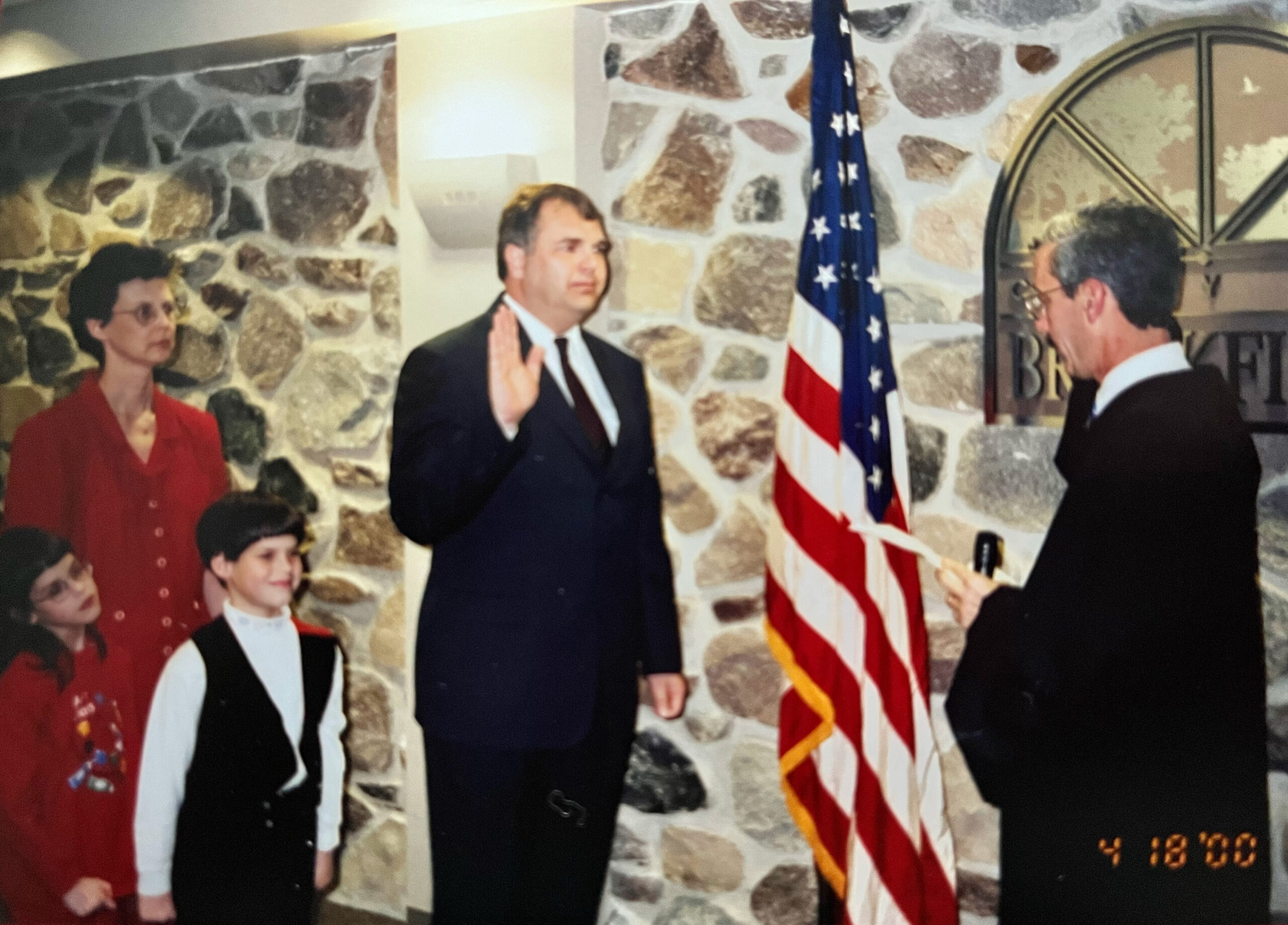 Man in a dark suit raises his right hand to take an oath as bystanders watch; American flag and stone wall behind.