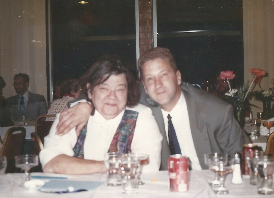 Two people pose for a photo at a restaurant table; the man has his arm around the older woman beside him, smiling softly.