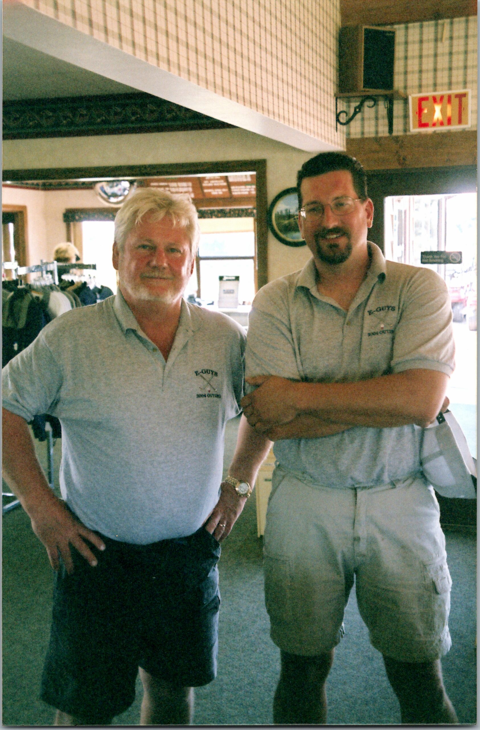 Two men pose together indoors, one with arms crossed, wearing light gray polo shirts and casual shorts/jeans; an exit sign is visible in the background.
