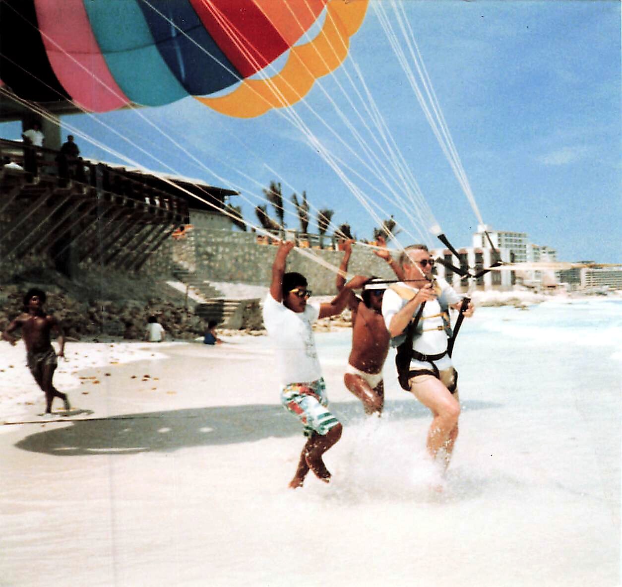 People on a sunny beach assist a parasail rider as a colorful parachute lifts into the sky over the water.