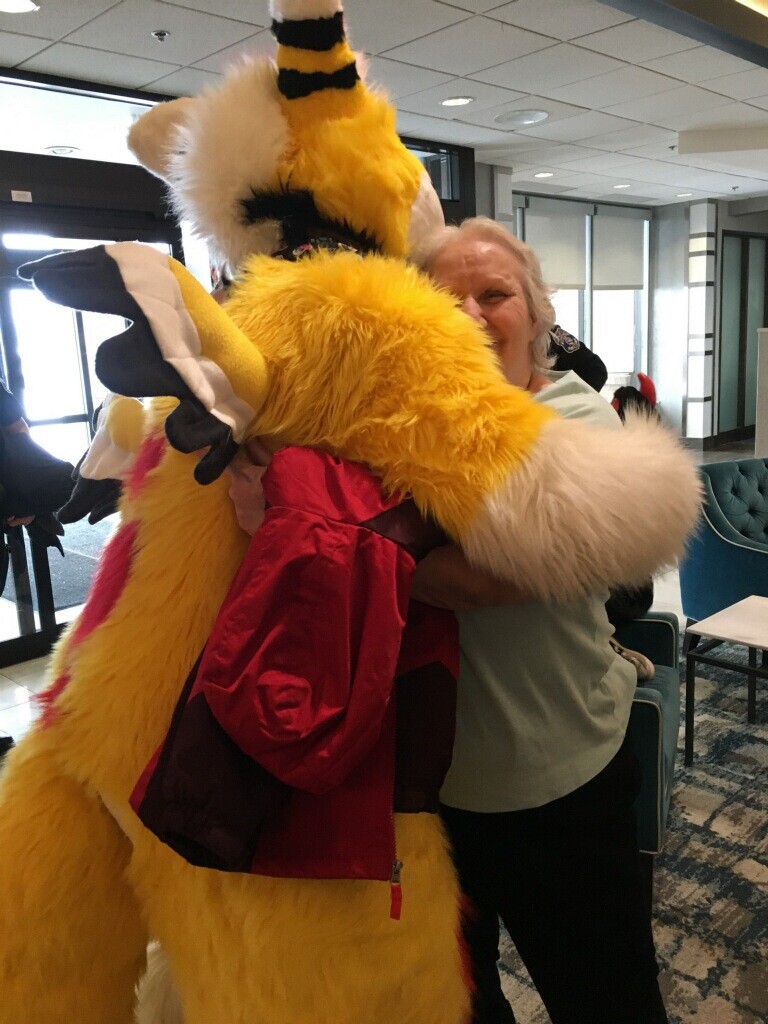 Person in a bright yellow furry dragon costume hugging a smiling woman indoors, with a red jacket draped over the costume's arm and seating area in the background.