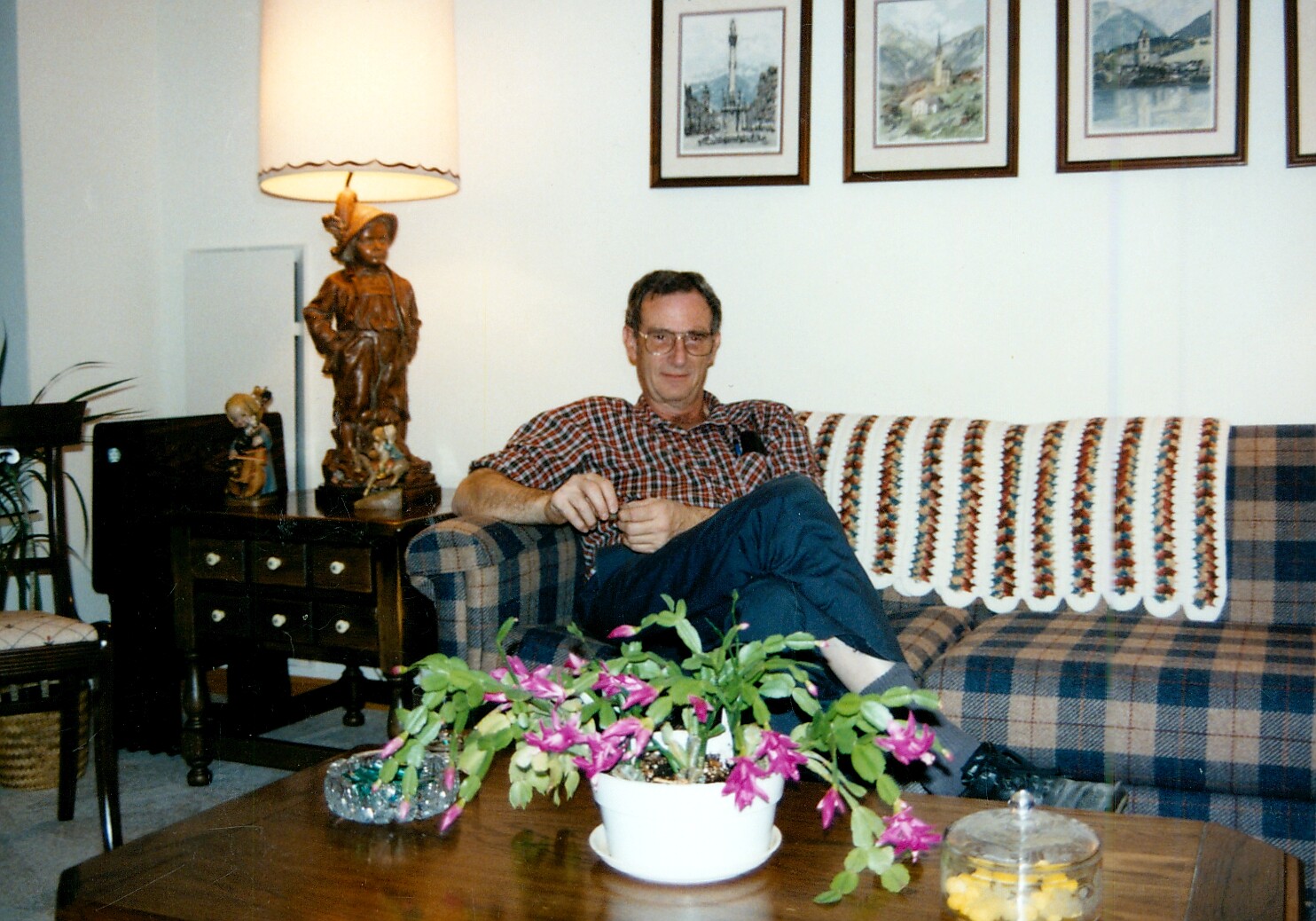 Man sits relaxed on a plaid sofa in a living room, with a crocheted throw and framed art on the wall behind him.