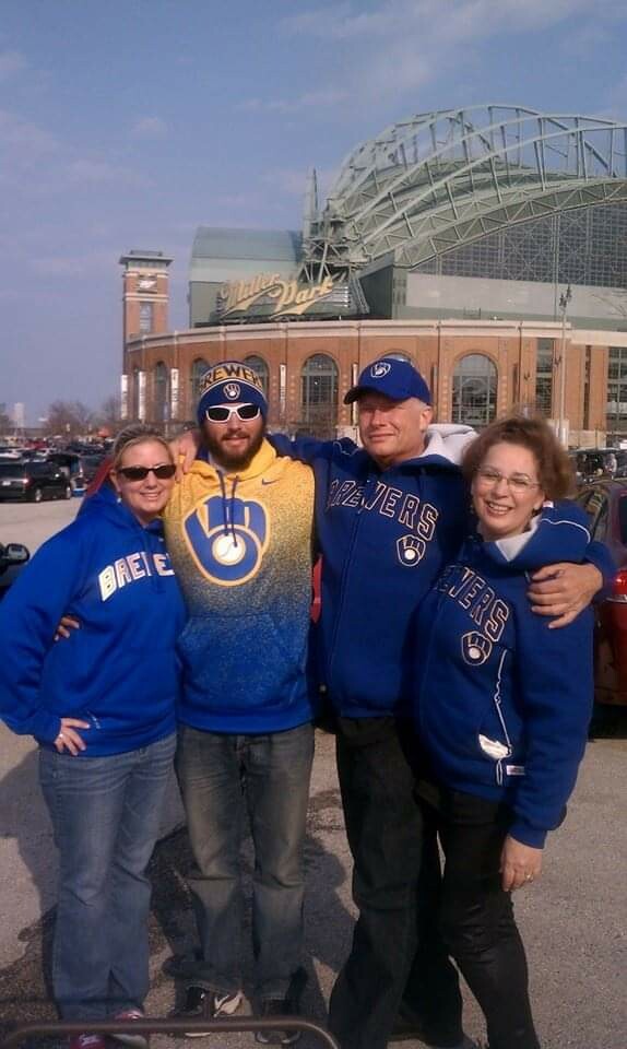 Four people wearing Milwaukee Brewers hoodies posing together outside a stadium on a sunny day.