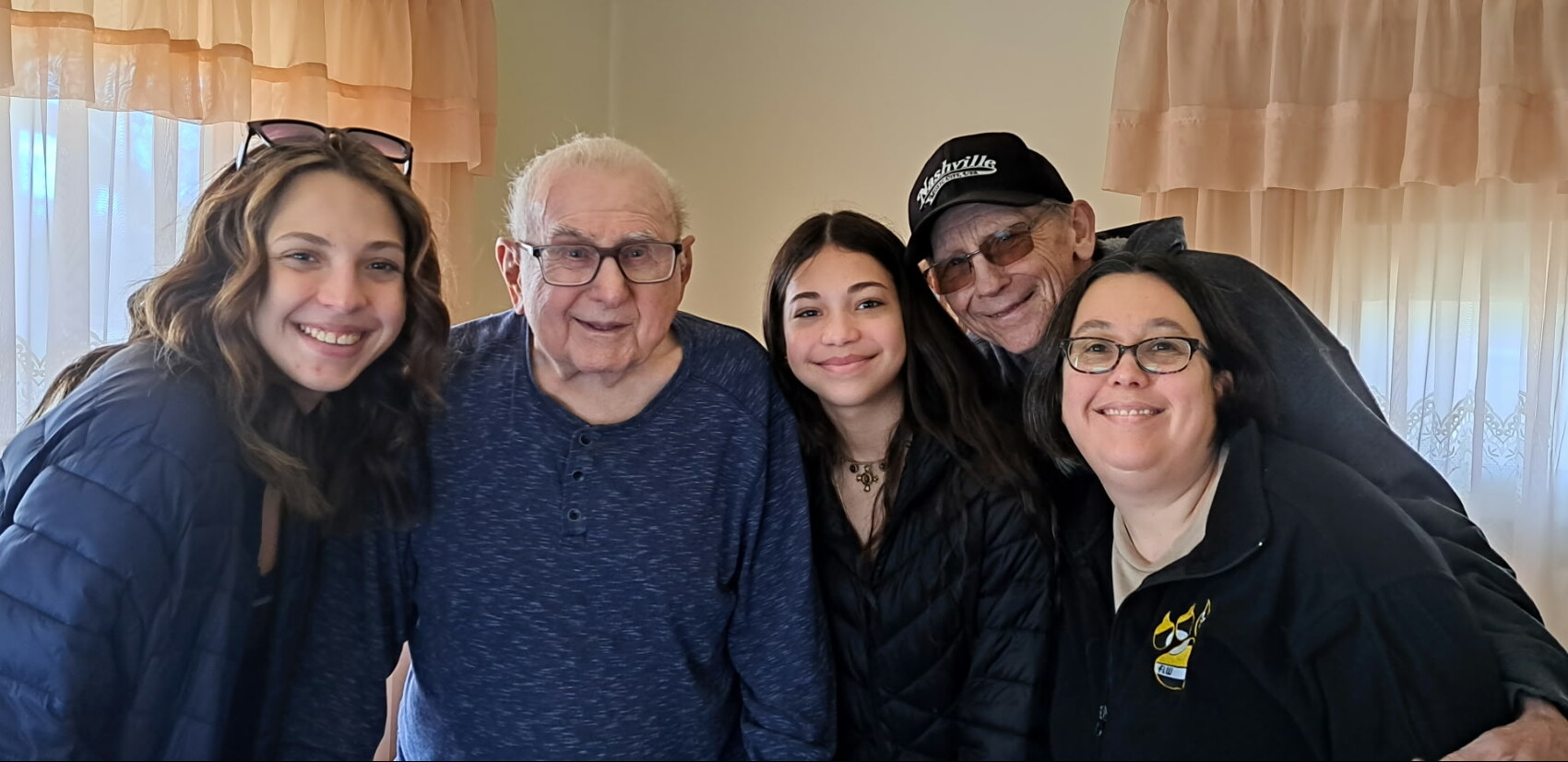 Five people of different ages posing together indoors, smiling at the camera with beige curtains in the background.