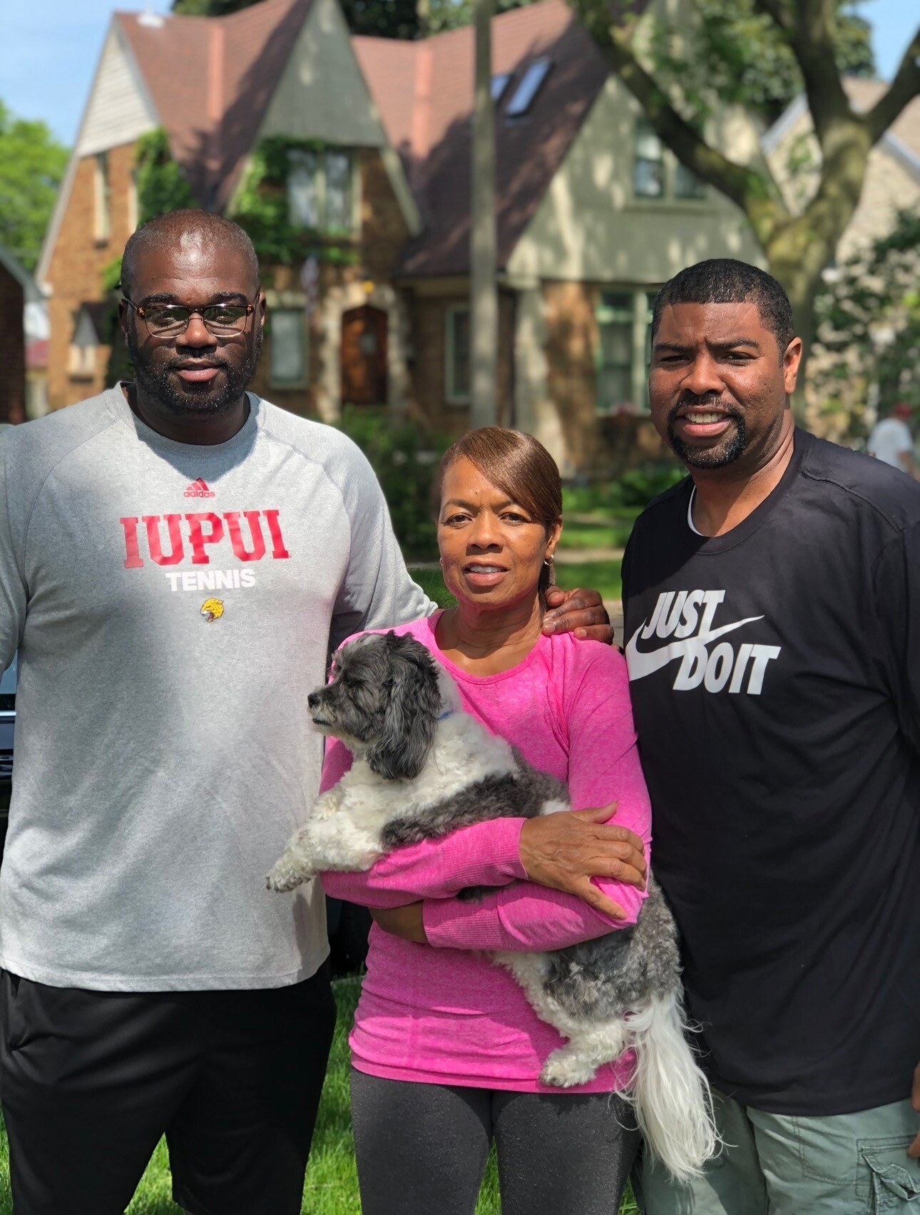 Three adults pose together outdoors in a sunny suburban yard, with a small fluffy dog held by the woman in pink center.