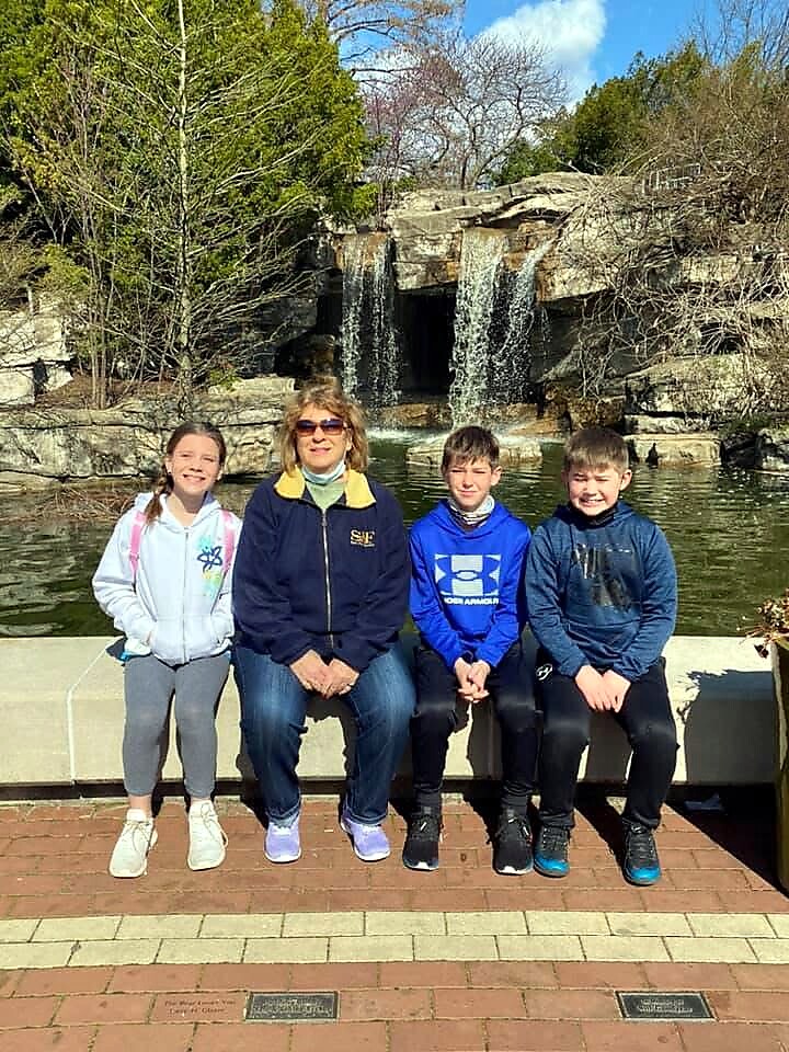 A woman and three kids sit on a brick ledge by a small waterfall in a park, posing for a photo.