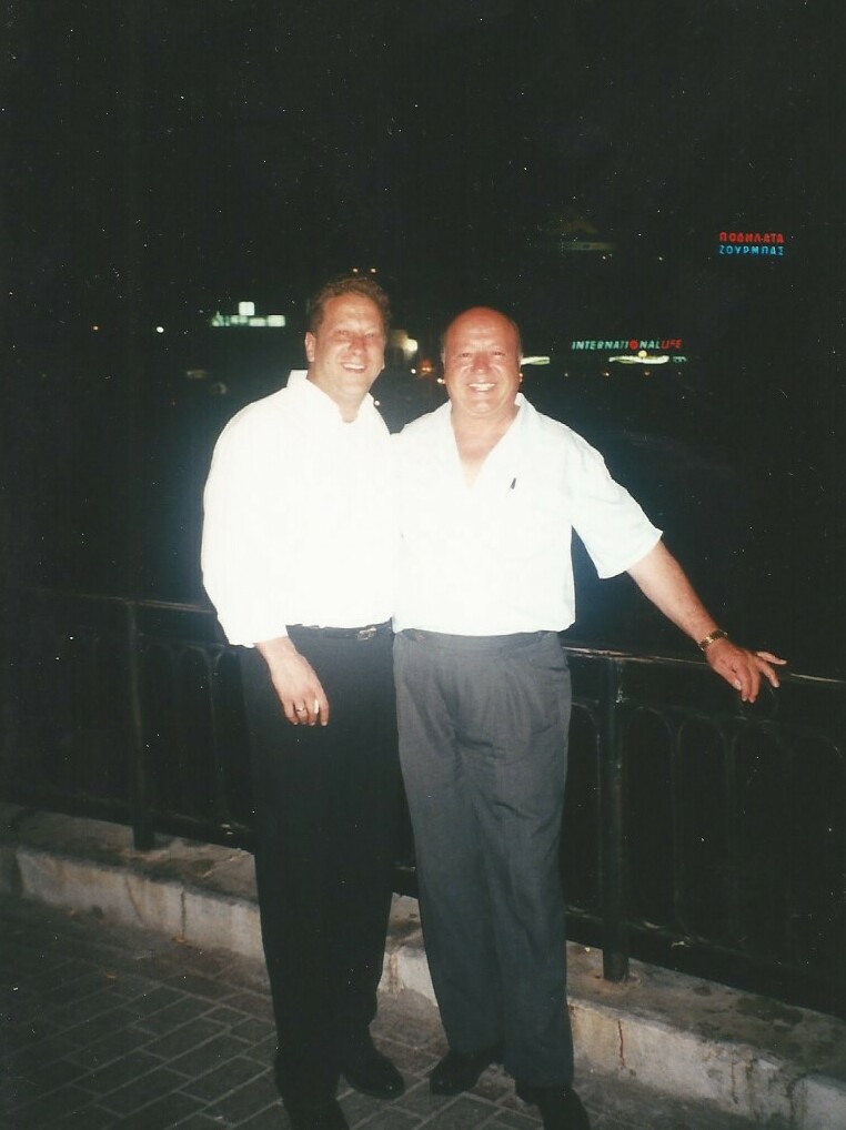 Two men in white shirts posing together at night by a railing, smiling with city lights behind them.