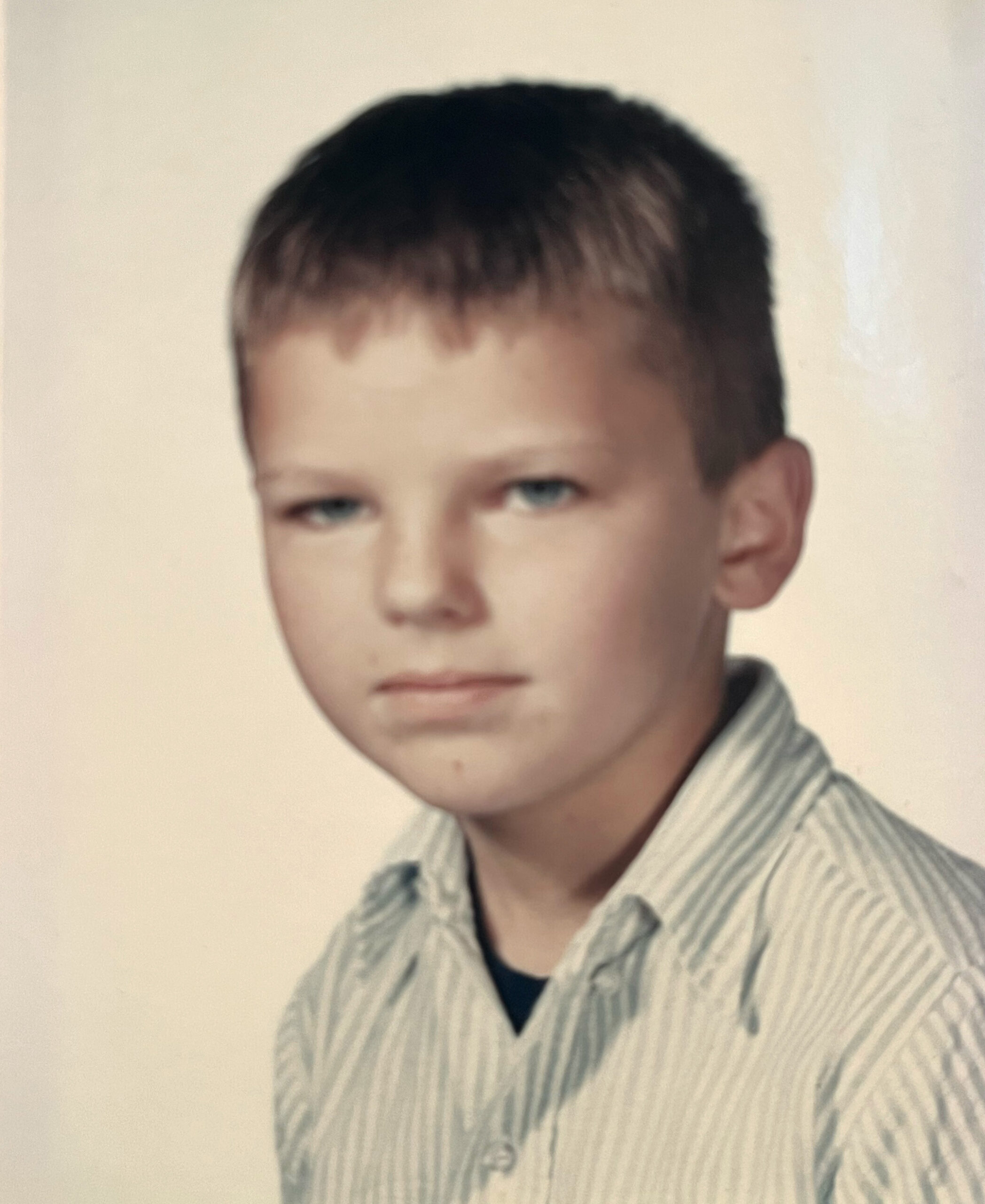 Portrait of a young boy with short dark hair wearing a light striped shirt, posed against a pale backdrop.