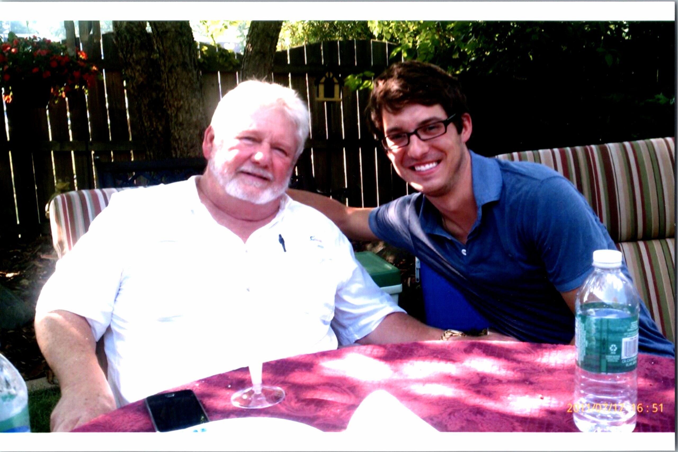 Two men pose for a photo at an outdoor table, smiling, with a wooden fence and greenery in the background.