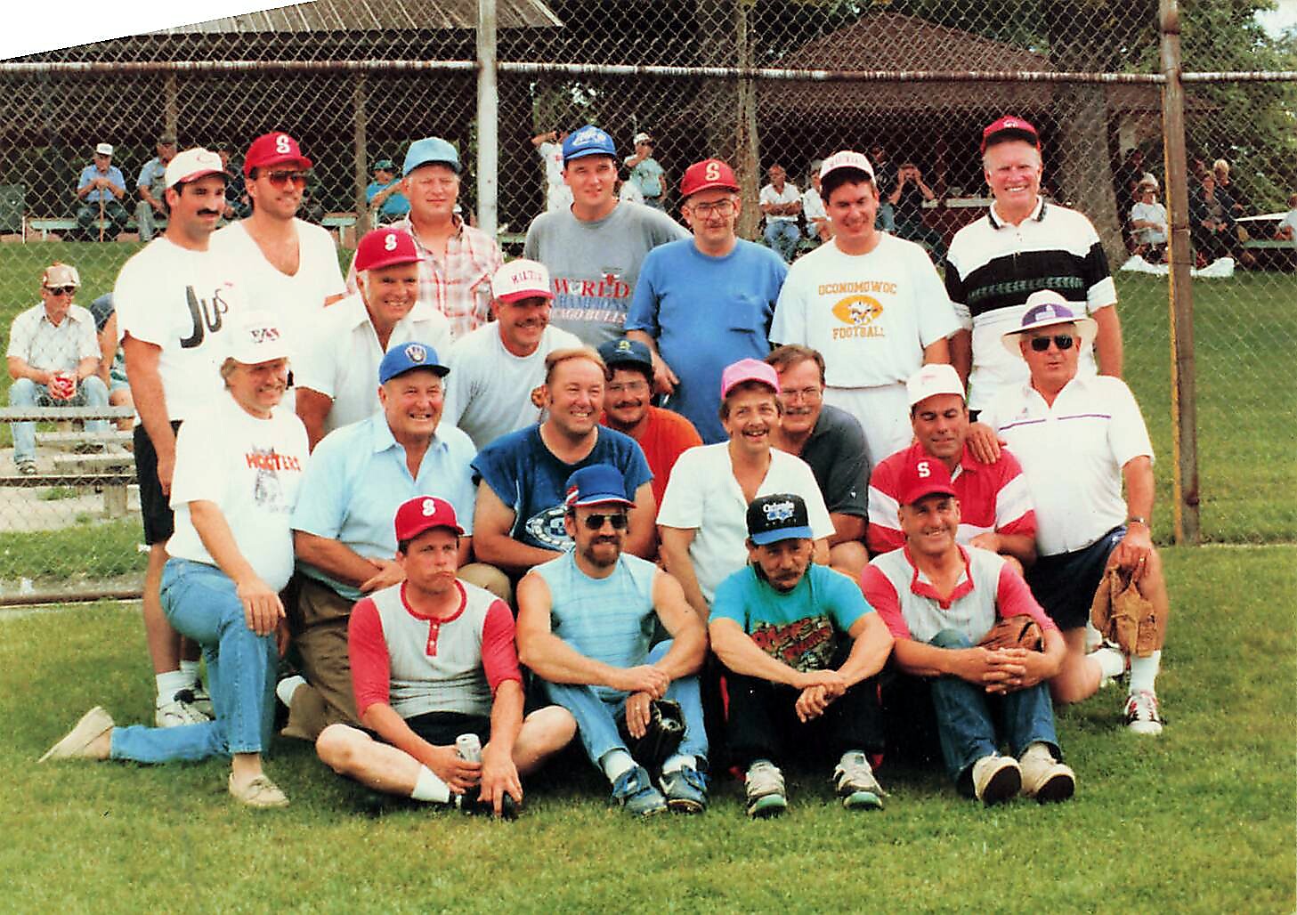 A large group of men in baseball caps and casual sportswear posing for a team photo on a grassy field behind a chain-link fence.
