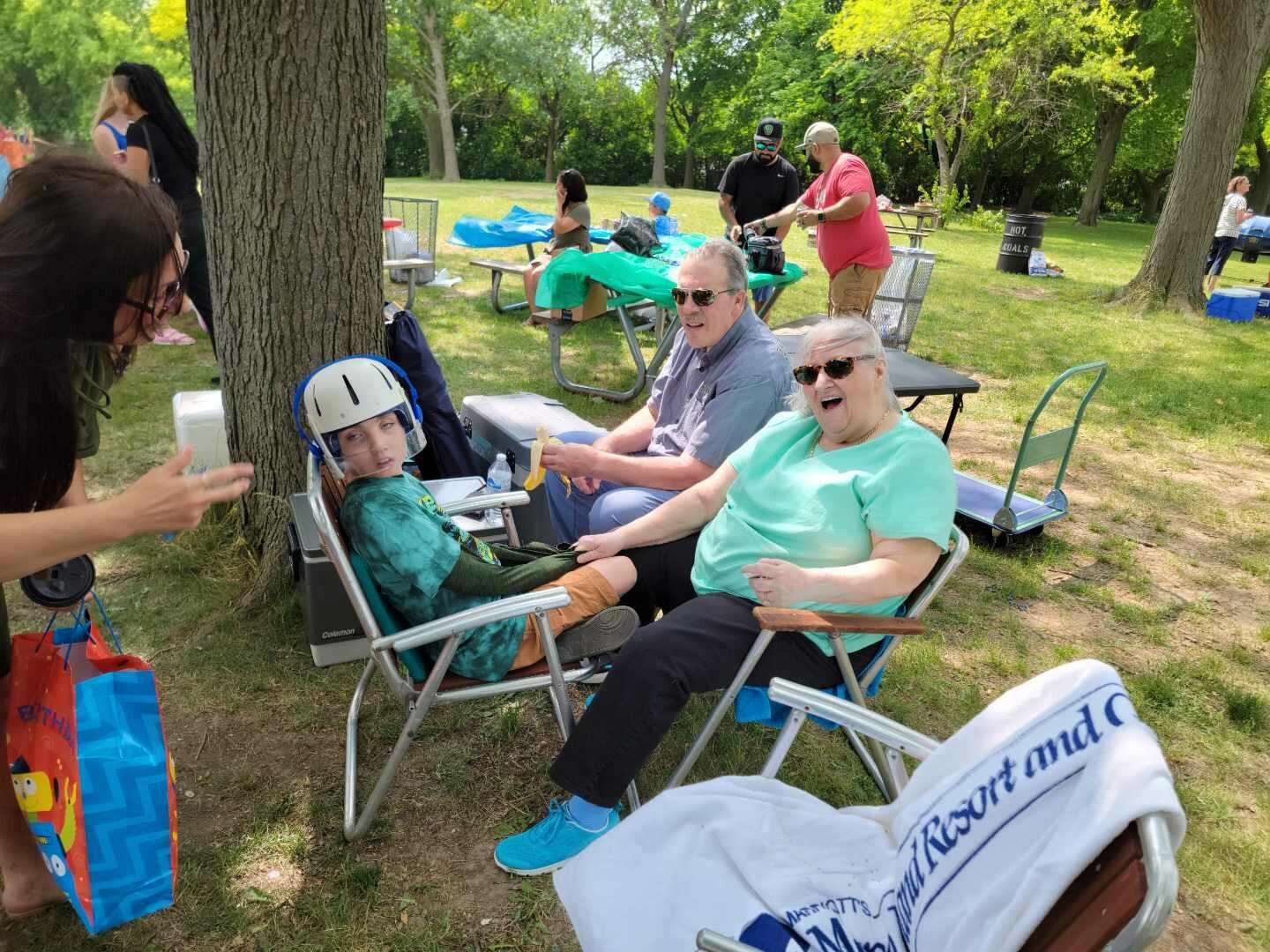 Child wearing a helmet sits in a folding chair at a park picnic, with smiling adults nearby under a tree.