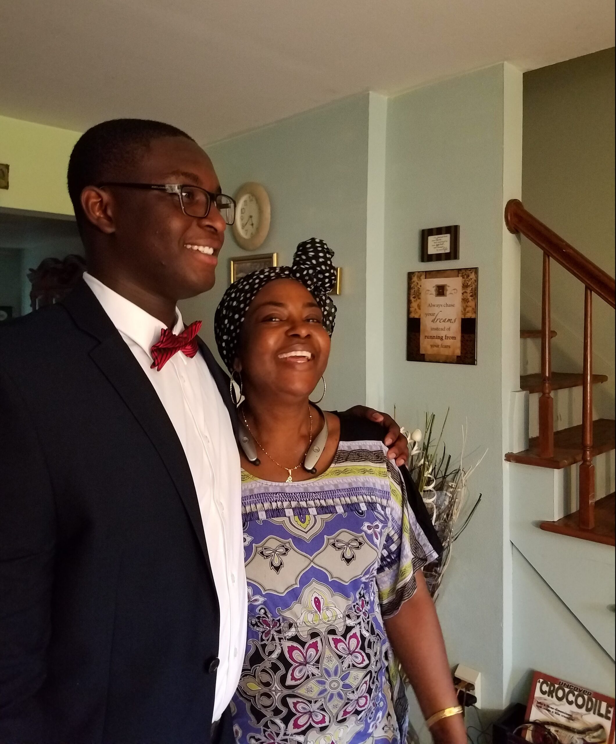 Smiling couple posing indoors: man in a suit with a red bow tie and glasses, woman in a colorful patterned top with a black-and-white head wrap, background includes wall art and a staircase.