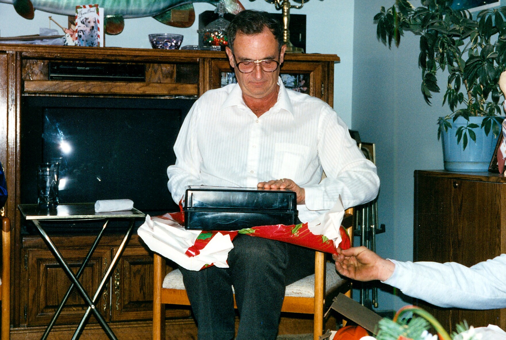 Man in a white shirt sits on a chair, opening a black rectangular gift box with torn wrapping paper on his lap in a living room setting.