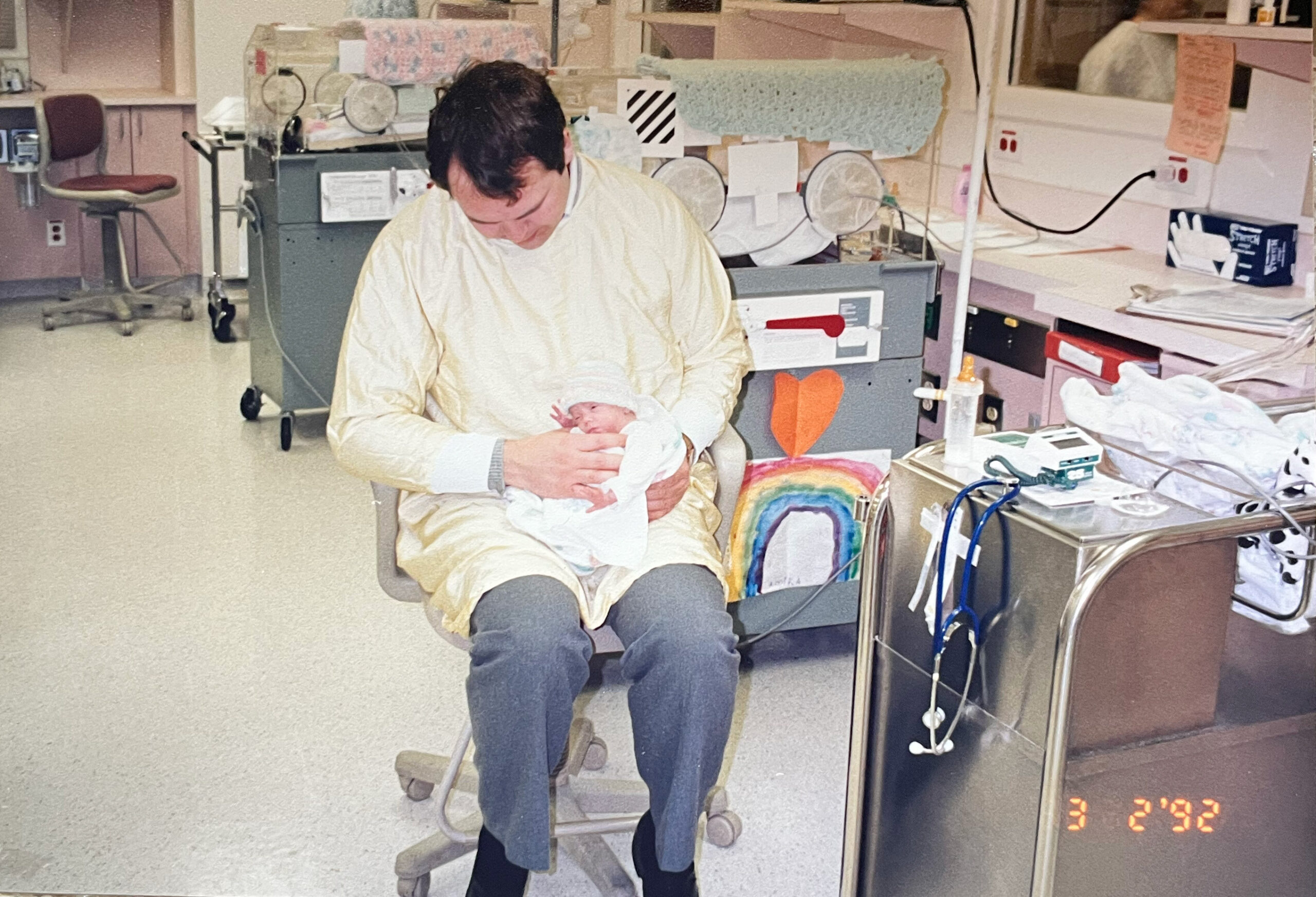Man in a hospital gown cradles a newborn swaddled in white in a maternity room with medical equipment around him.
