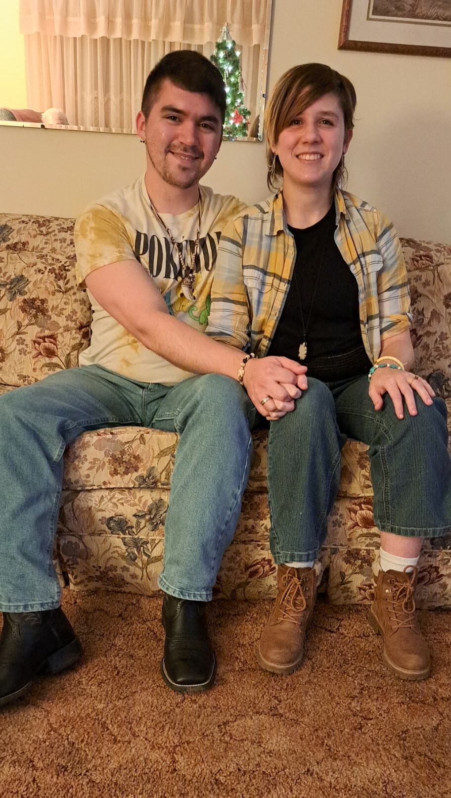 Couple sitting closely on a floral sofa, holding hands and smiling, with a Christmas tree in the background.