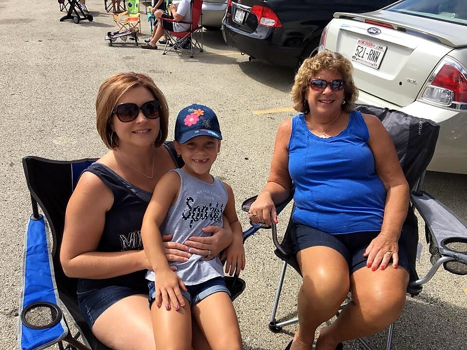 Woman wearing sunglasses sits next to a smiling young girl on her lap, with an older woman in a blue tank top beside them in a parking lot.