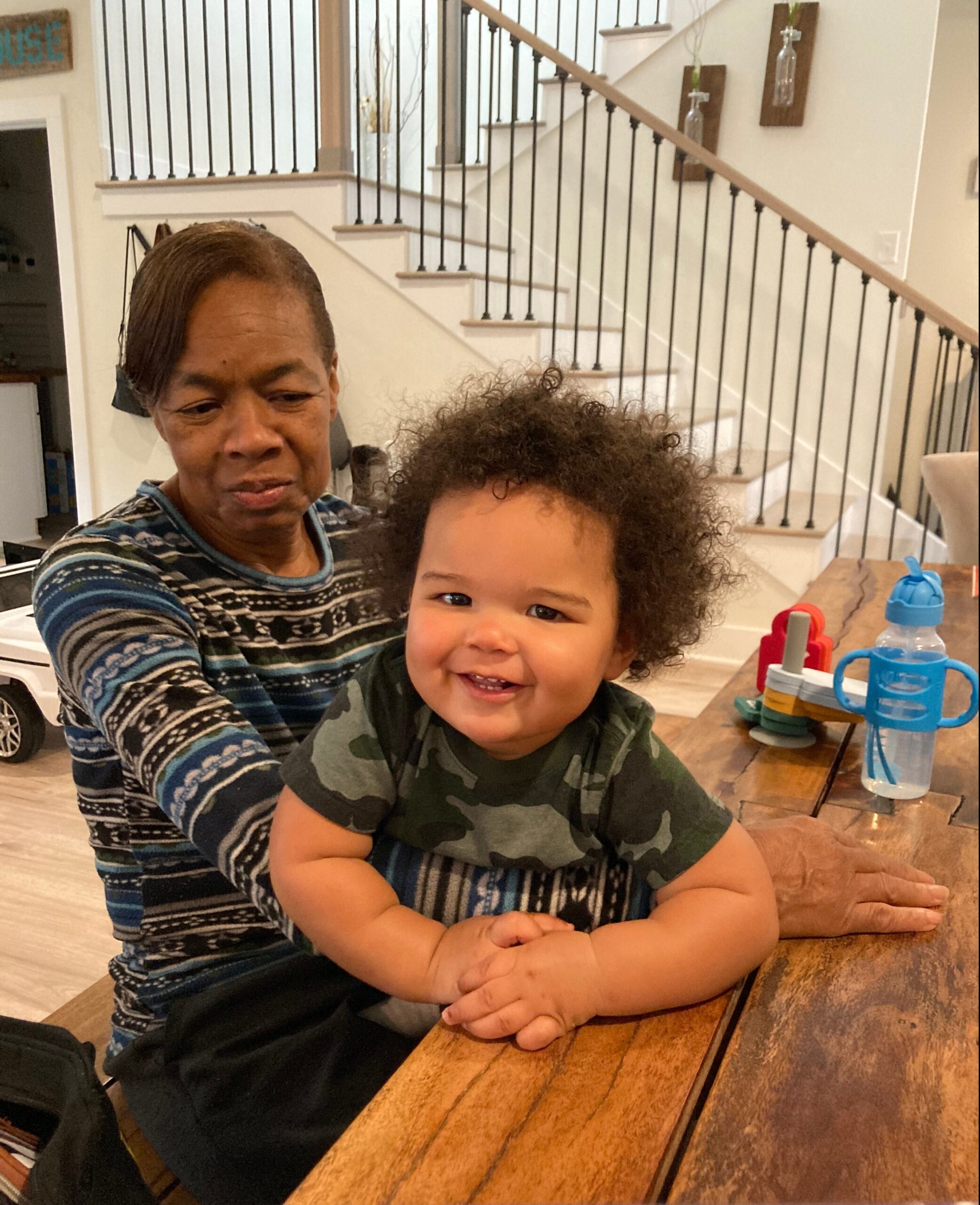 Woman and smiling toddler seated at a wooden table indoors, with a staircase and wall decor in the background.