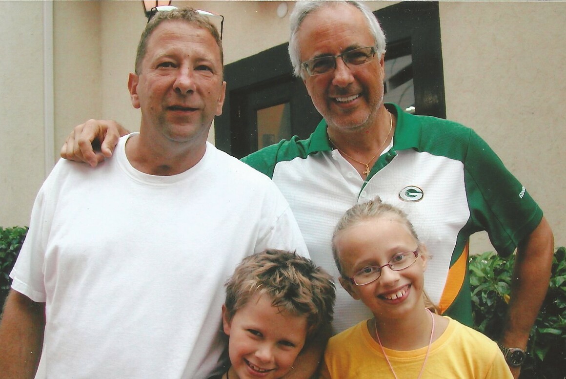 Four people posing outdoors: two adult men behind, with a boy and a girl in front, all smiling at the camera.