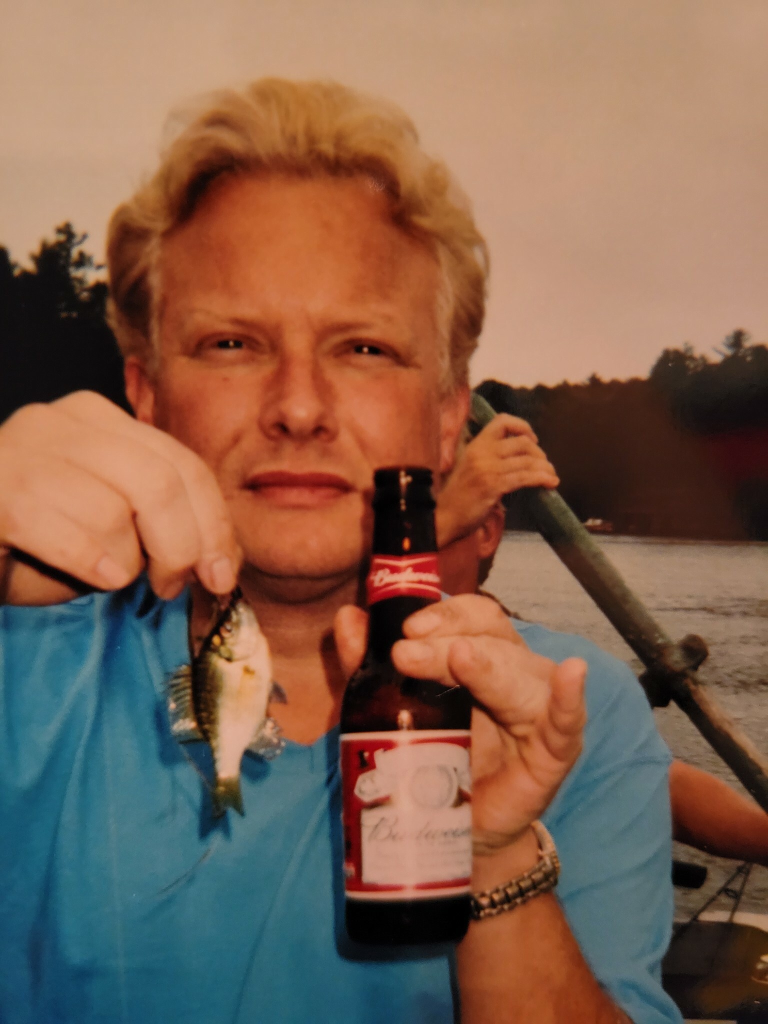 Man with blond hair in a blue shirt holding a Budweiser beer bottle on a boat by a lake at sunset.